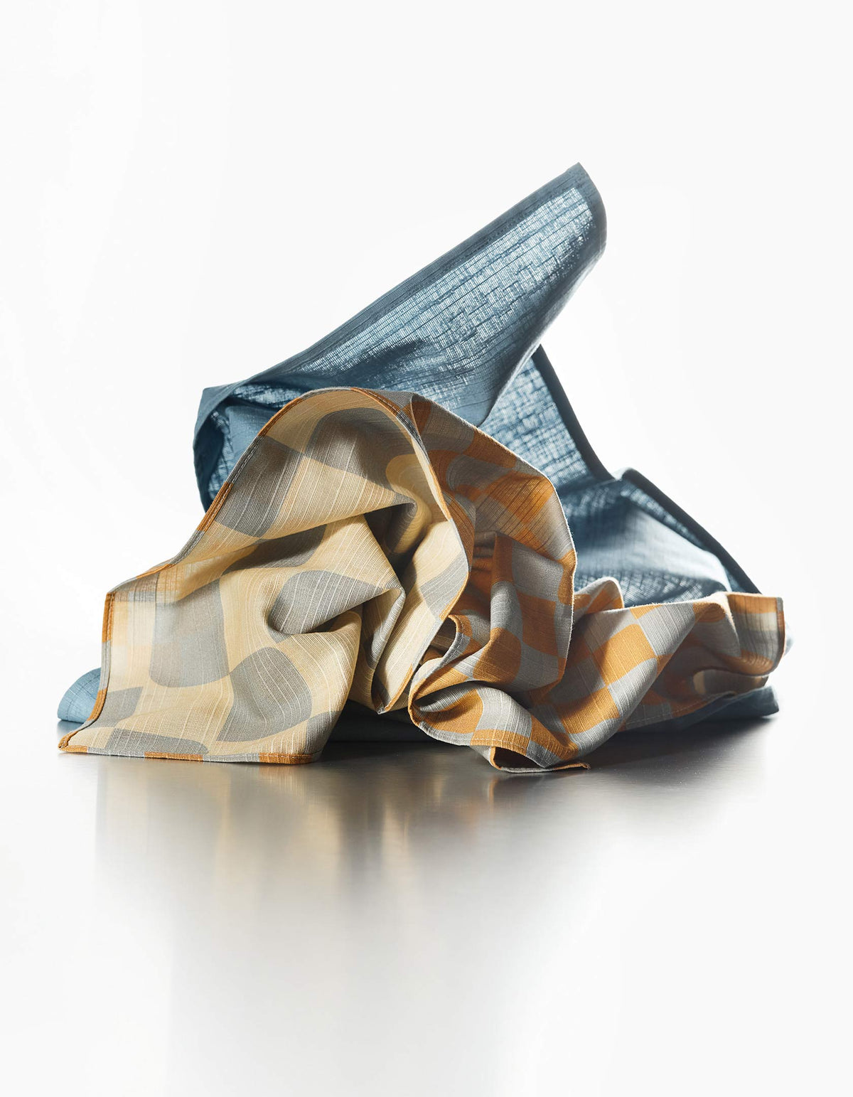 Tortuga Forma Dinner Napkins Double Sided Checker - Quiet Season gathered into loose folds on a light surface, revealing contrasting dark teal, cream, grey and mustard checker patterns.