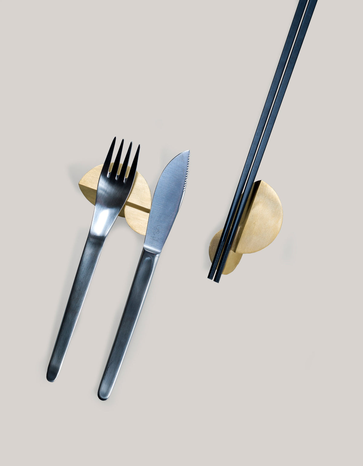 Overhead view of two Tortuga Forma Kaolin Brass Utensil Holders on a light grey surface, with a fork and knife resting across one and dark chopsticks across the other, showing their rounded gold-toned tops and central grooves.