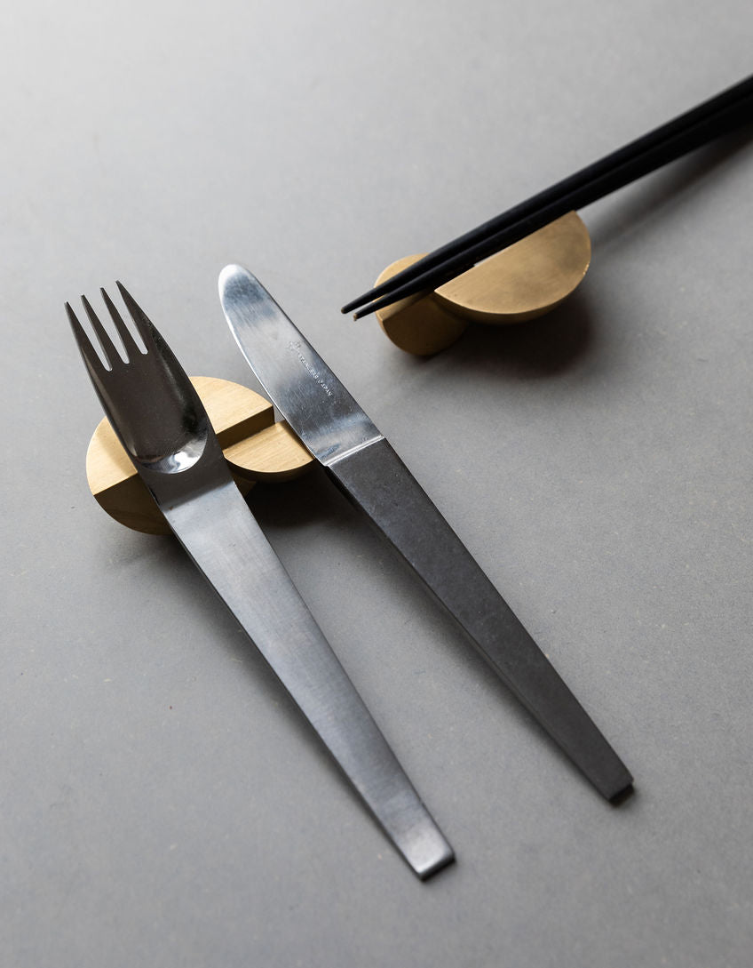 Two gold-toned Tortuga Forma Kaolin Brass Utensil Holders on a grey tabletop, one supporting a silver-coloured fork and knife and the other holding black chopsticks, showing curved, intersecting forms with shallow grooves across their tops.