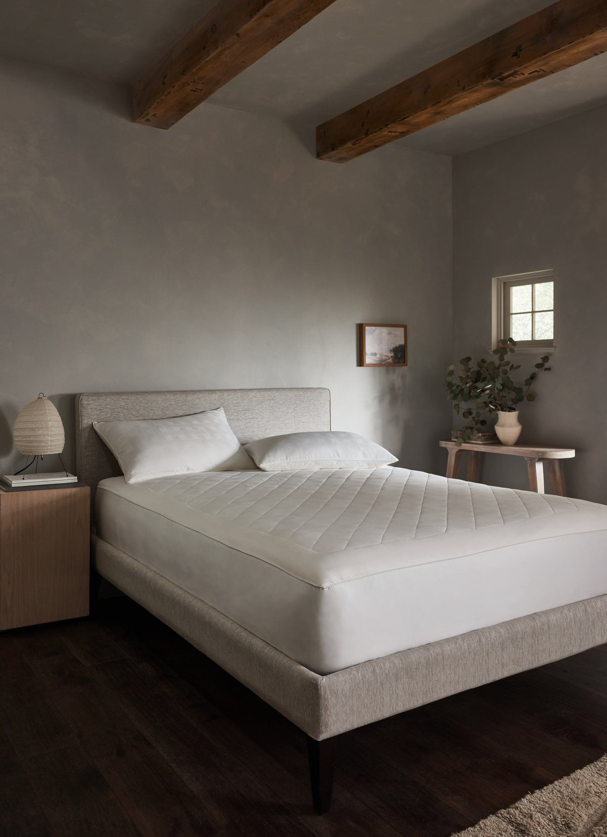 White mattress pad fitted over a bed in a bedroom with gray walls, exposed wooden ceiling beams, white pillows, and dark wood flooring.