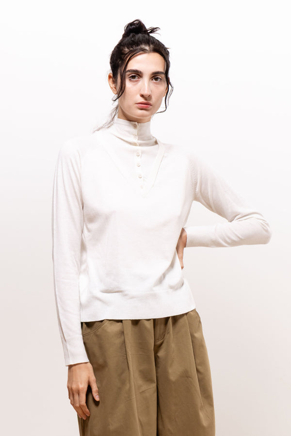 Model wearing the ivory Paulette Montagne Top with khaki-brown trousers, posing with one hand at her waist, showing its high neckline, short button placket, long sleeves, and gently fitted shape.