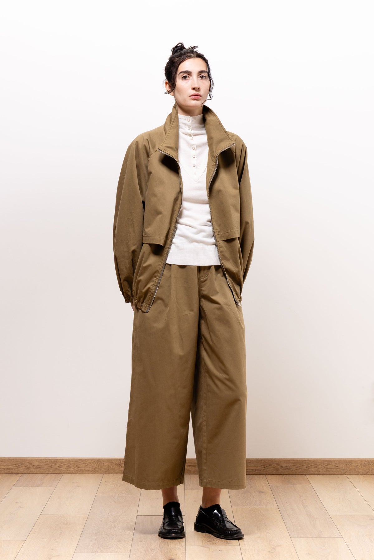 Model wearing khaki-brown Paulette Lee Trousers with a matching open bomber jacket, ivory top, and black shoes, standing with hands in pockets to show the relaxed fit and cropped wide legs.