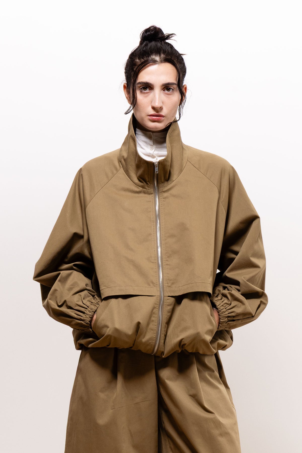 Front view of a model wearing the khaki-brown Paulette Lee Bomber Jacket fully zipped over a white high-neck top, with both hands in the lower pockets and matching trousers.