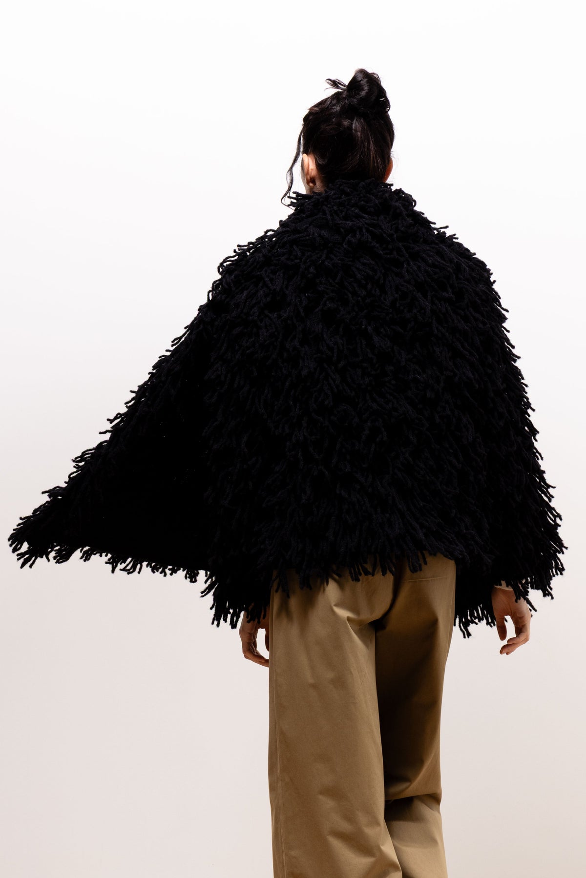 Back view of a model wearing the black Paulette Saint-Jean-de-Luz Oversize Scarf across her shoulders, showing its broad coverage and uneven strand-covered edge above khaki-brown trousers.