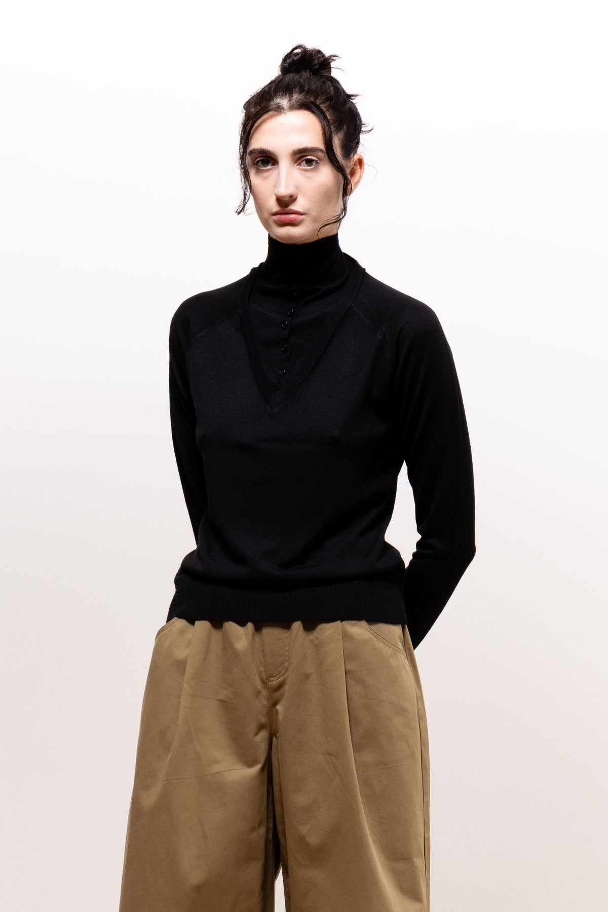 Model facing forward in the black Paulette Montagne Top and khaki-brown trousers, with her hands behind her back, showing the high neckline and softly draped front.