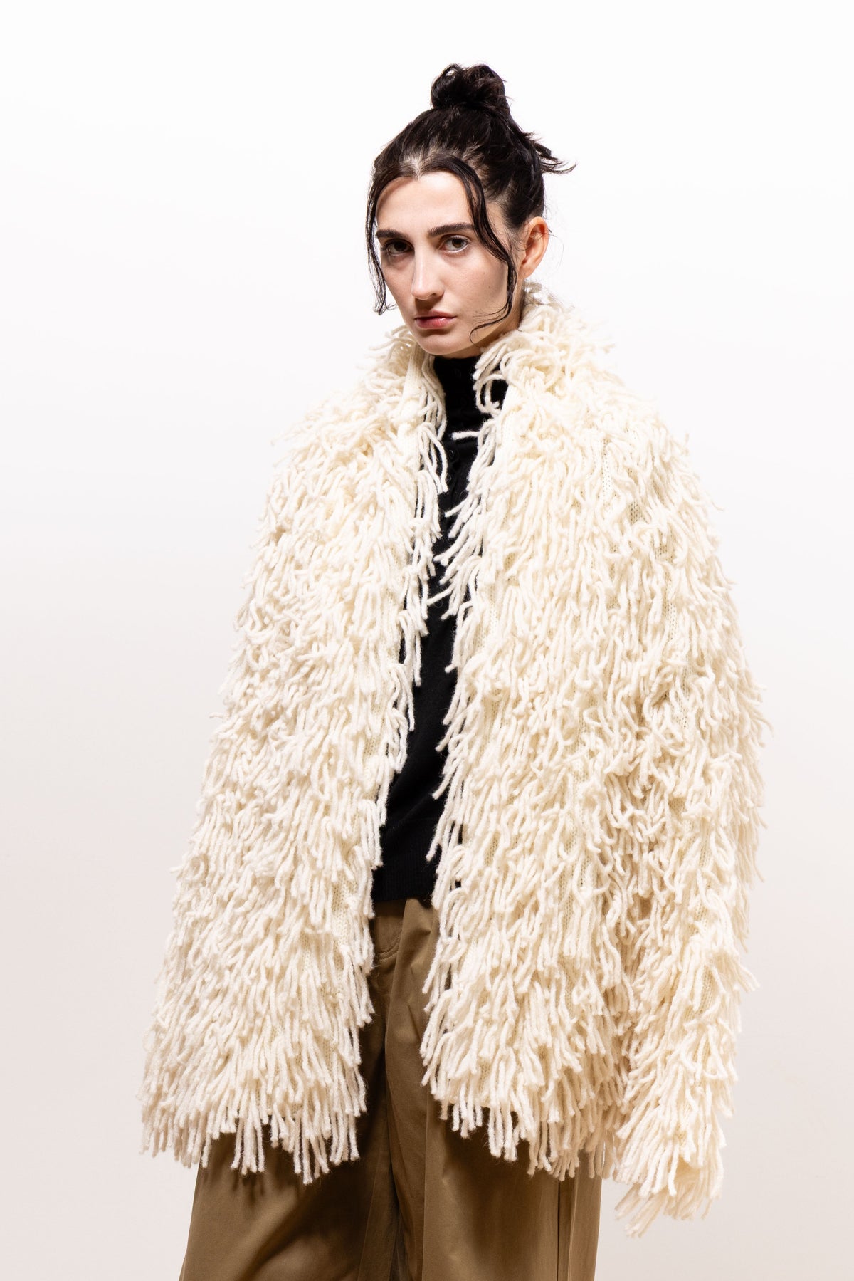 Model wearing the ivory Paulette Saint-Jean-de-Luz Oversize Scarf draped around her neck and shoulders, with long shaggy strands covering her arms and hanging to her upper thighs over a black top and khaki-brown trousers.
