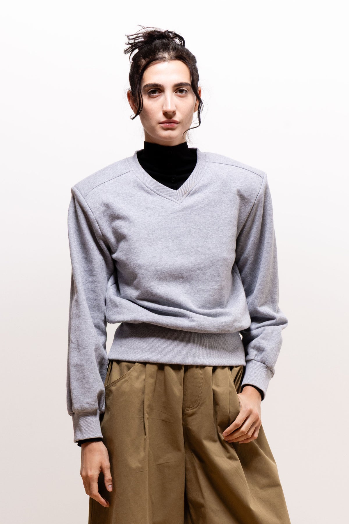 Model wearing the light gray Paulette Madeleine Top over a black high-neck layer and khaki-brown trousers, showing soft folds through the body and sleeves above the fitted waistband.