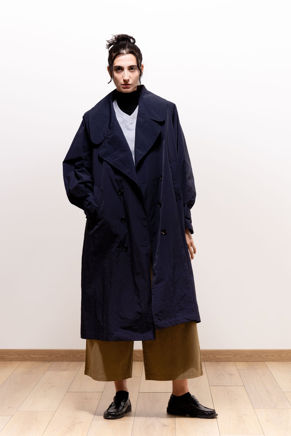 Model wearing the dark navy Paulette Louise Trench Coat unbelted with one hand in a pocket, showing its loose knee-length silhouette and broad lapels over layered tops and khaki-brown trousers.