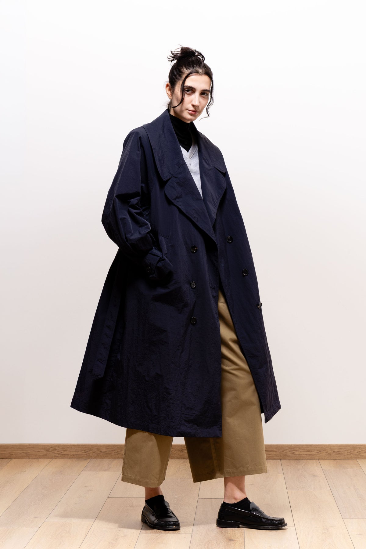 Three-quarter view of a model wearing the dark navy Paulette Louise Trench Coat open with one hand in a pocket, showing the flared hem and khaki-brown trousers beneath.