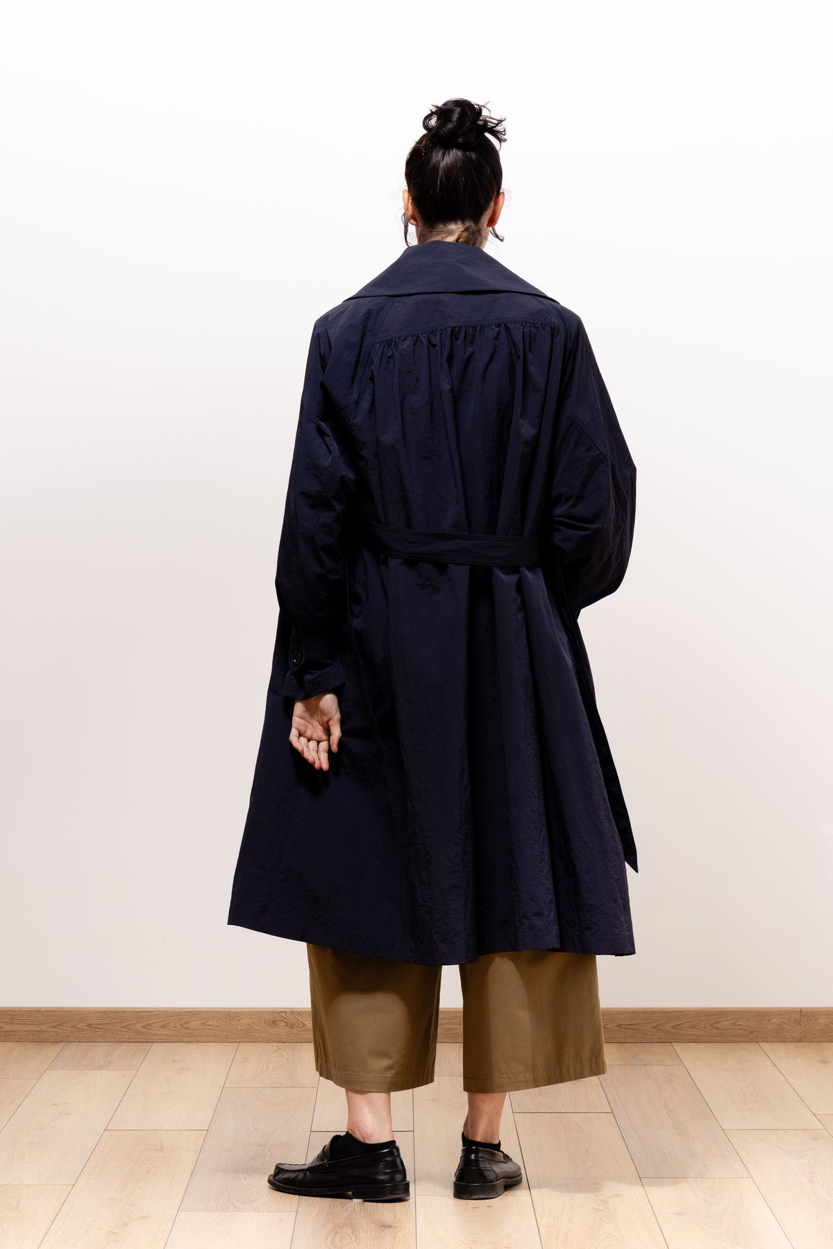 Back view of a model wearing the dark navy Paulette Louise Trench Coat, showing gathers beneath the shoulder yoke, a belt around the waist, and a full skirt falling to the knees.