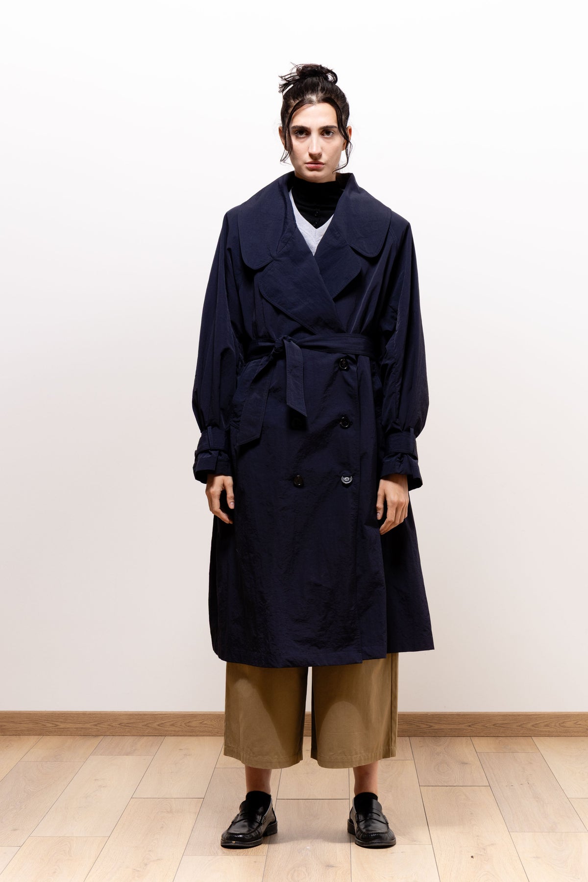 Front view of a model wearing the dark navy Paulette Louise Trench Coat belted at the waist over a black high-neck top and cropped khaki-brown trousers, paired with black shoes.