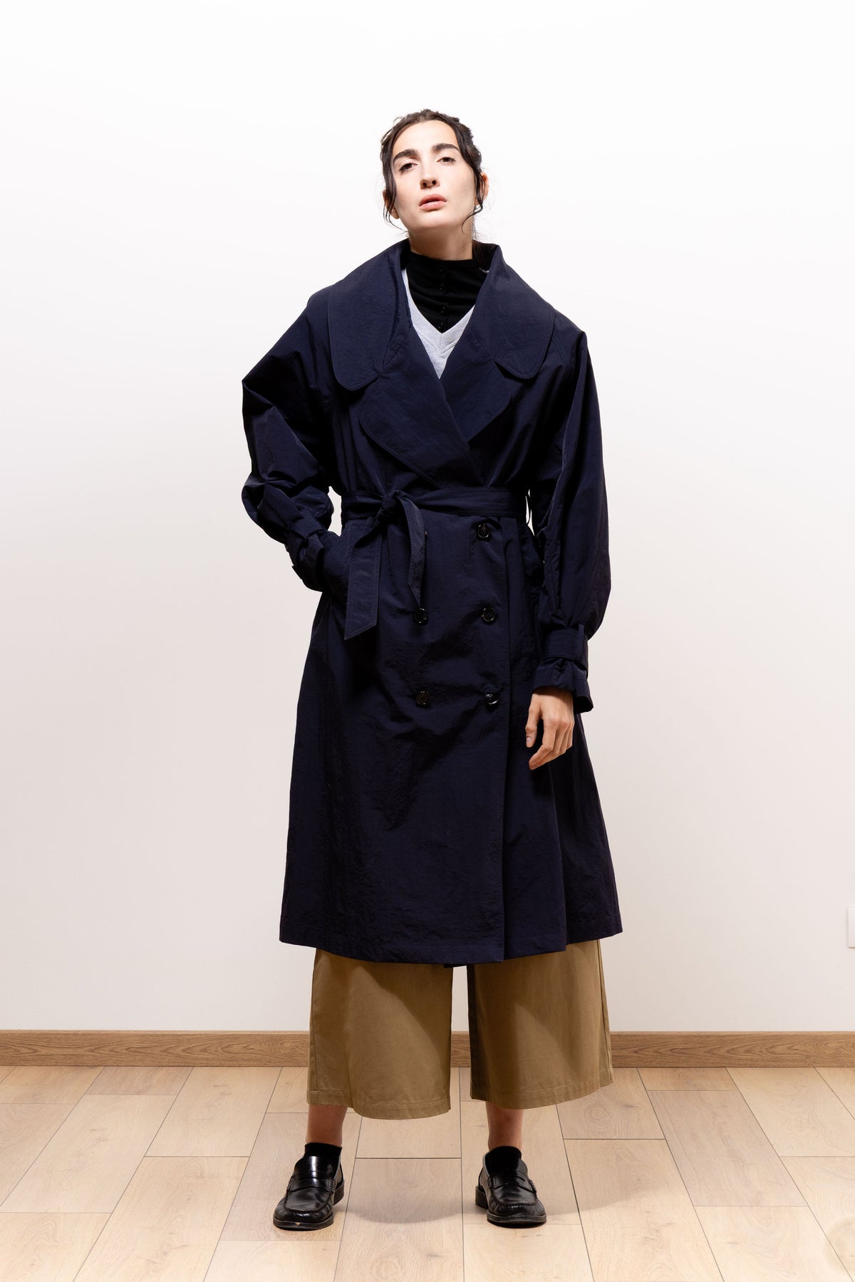 Model facing forward with her chin raised in the dark navy Paulette Louise Trench Coat, its belt tied at the waist and one hand resting in a pocket.