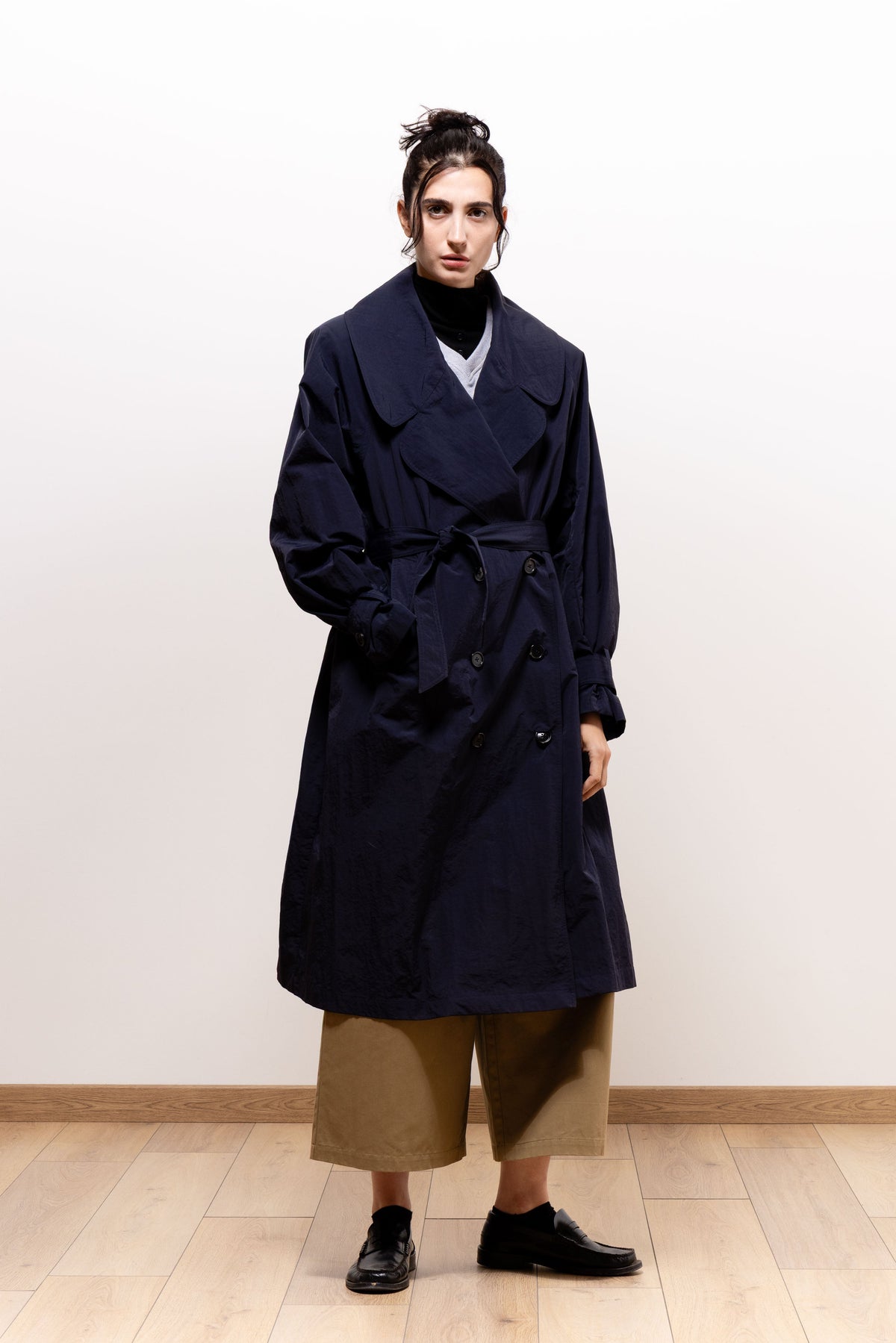 Front view of a model wearing the dark navy Paulette Louise Trench Coat with both hands in its pockets, showing the tied waist belt, overlapping lapels, and knee-length hem.