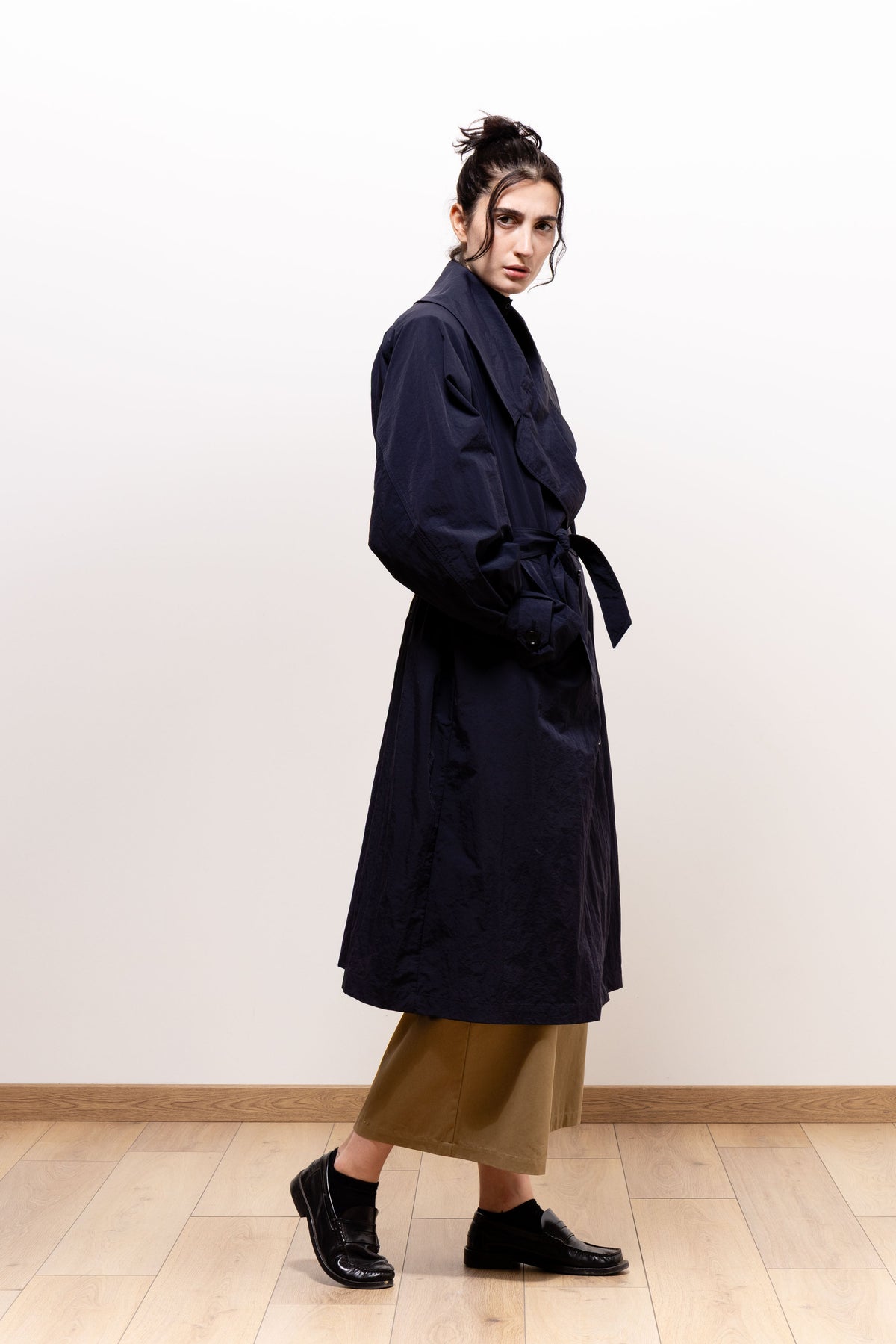 Side view of a model looking toward the camera in the belted dark navy Paulette Louise Trench Coat, showing its gathered waist and flared skirt over cropped khaki-brown trousers and black shoes.
