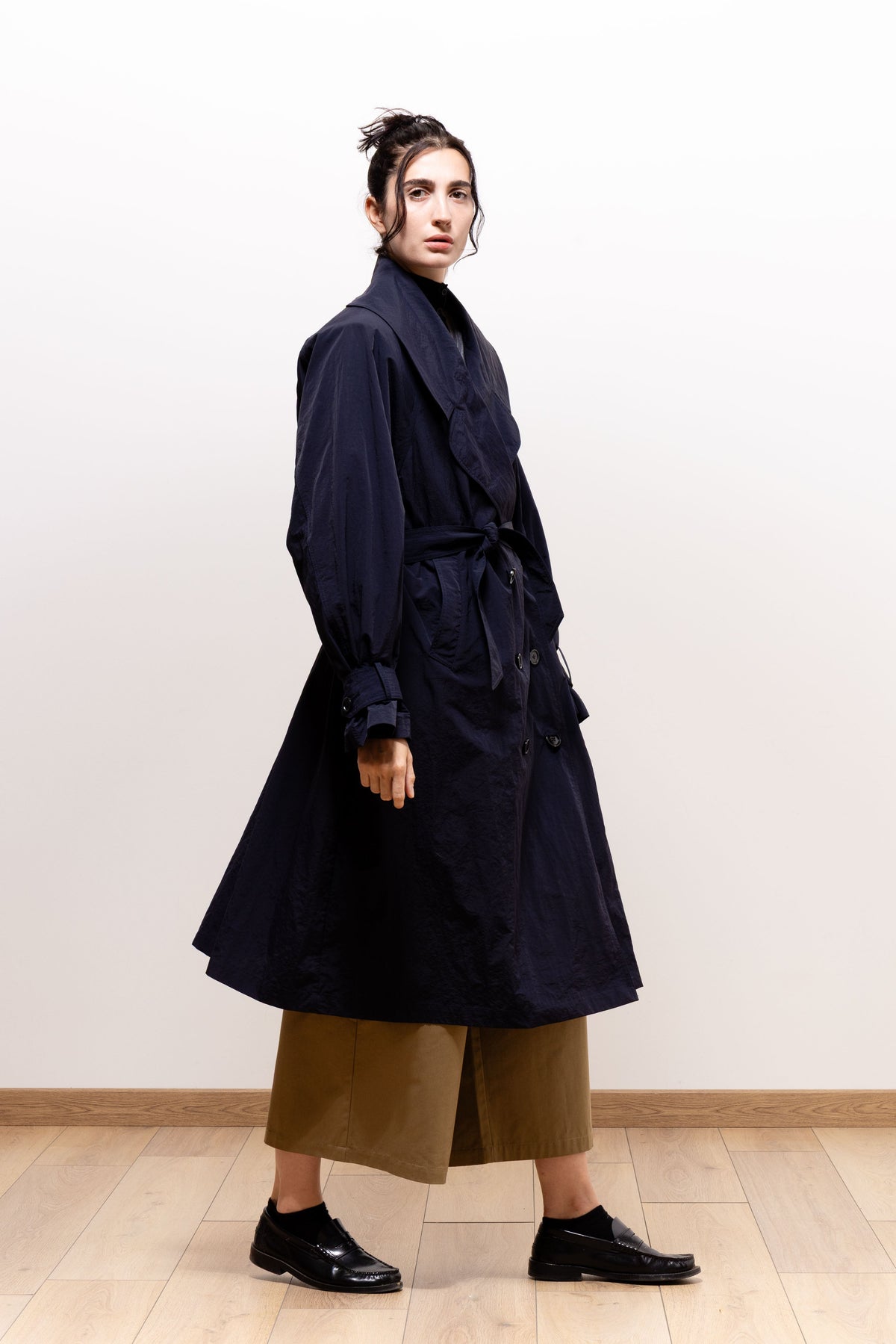 Three-quarter view of a model wearing a belted navy trench coat over cropped khaki-brown trousers and black shoes; the Paulette Louise Tier Skirt is not visible.