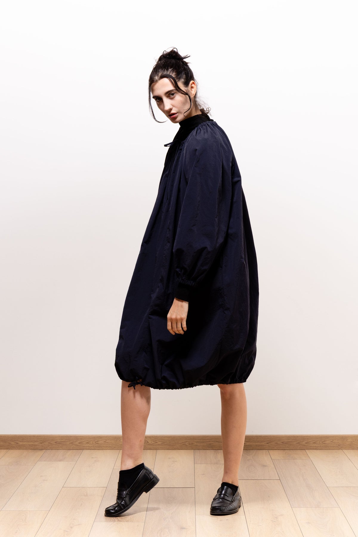 Three-quarter side view of a model wearing the dark navy Paulette Georgia Dress, showing the billowing body and gathered hem, with one foot extended on a pale wooden floor.
