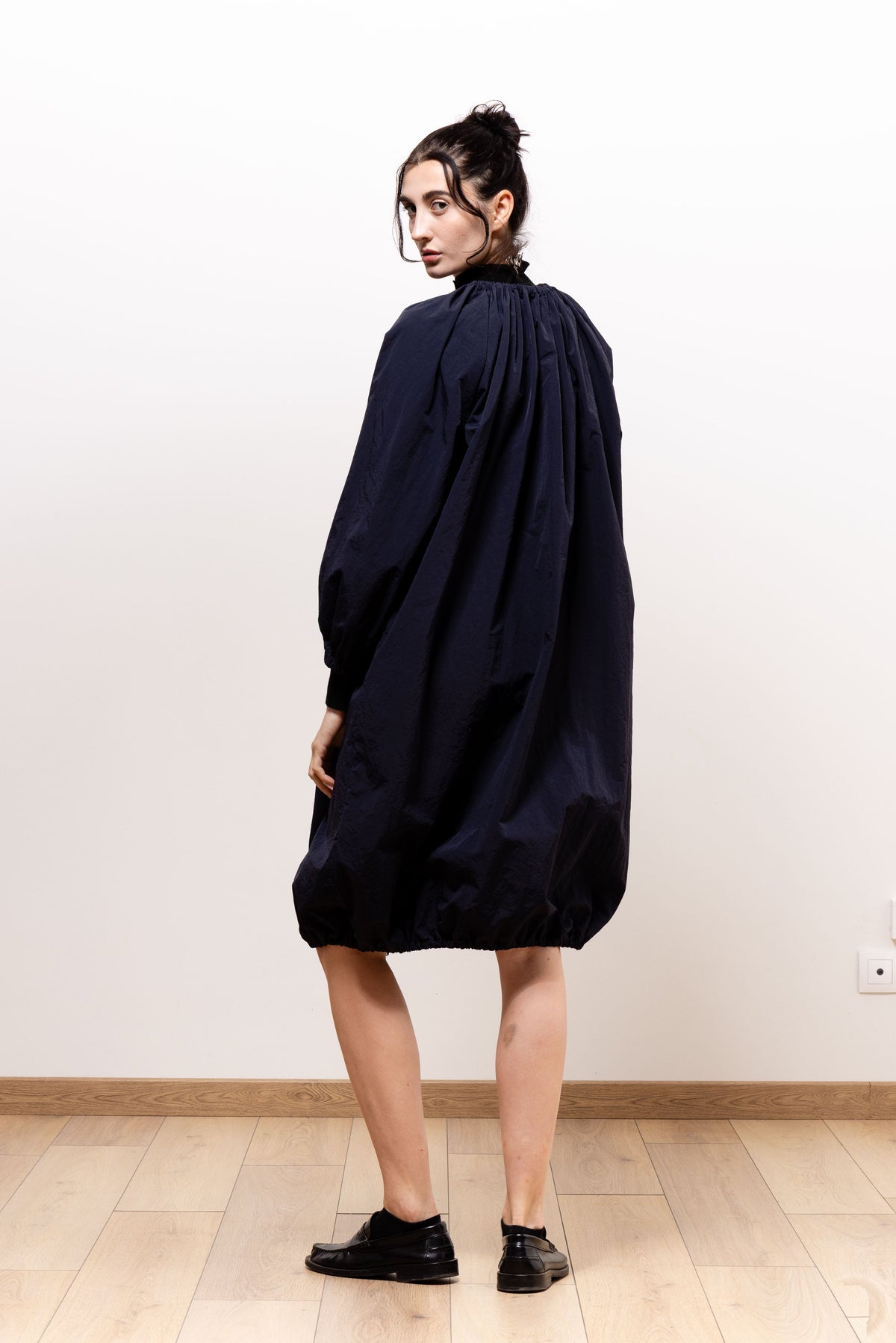 Three-quarter back view of a model wearing the dark navy Paulette Georgia Dress and black shoes, looking over her shoulder to show the upper-back gathers and full skirt narrowing into a bubble hem.