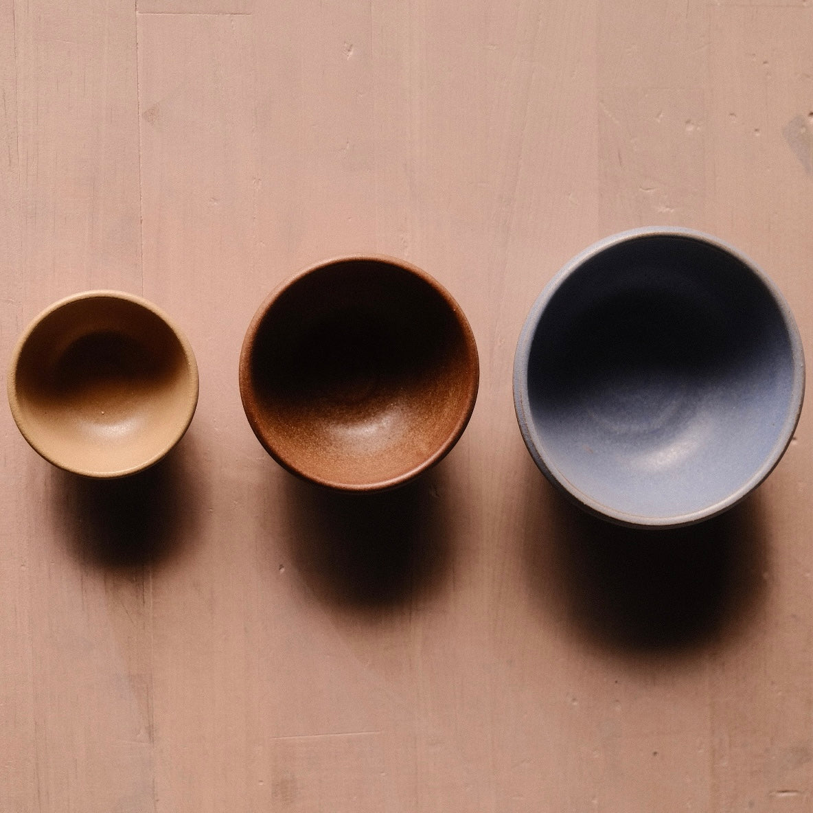 Overhead view of three bowls accompanying the Settle Ceramics Wander Pinch Bowl, arranged from small golden tan to medium reddish brown and larger grey, with rounded interiors and shadows below.
