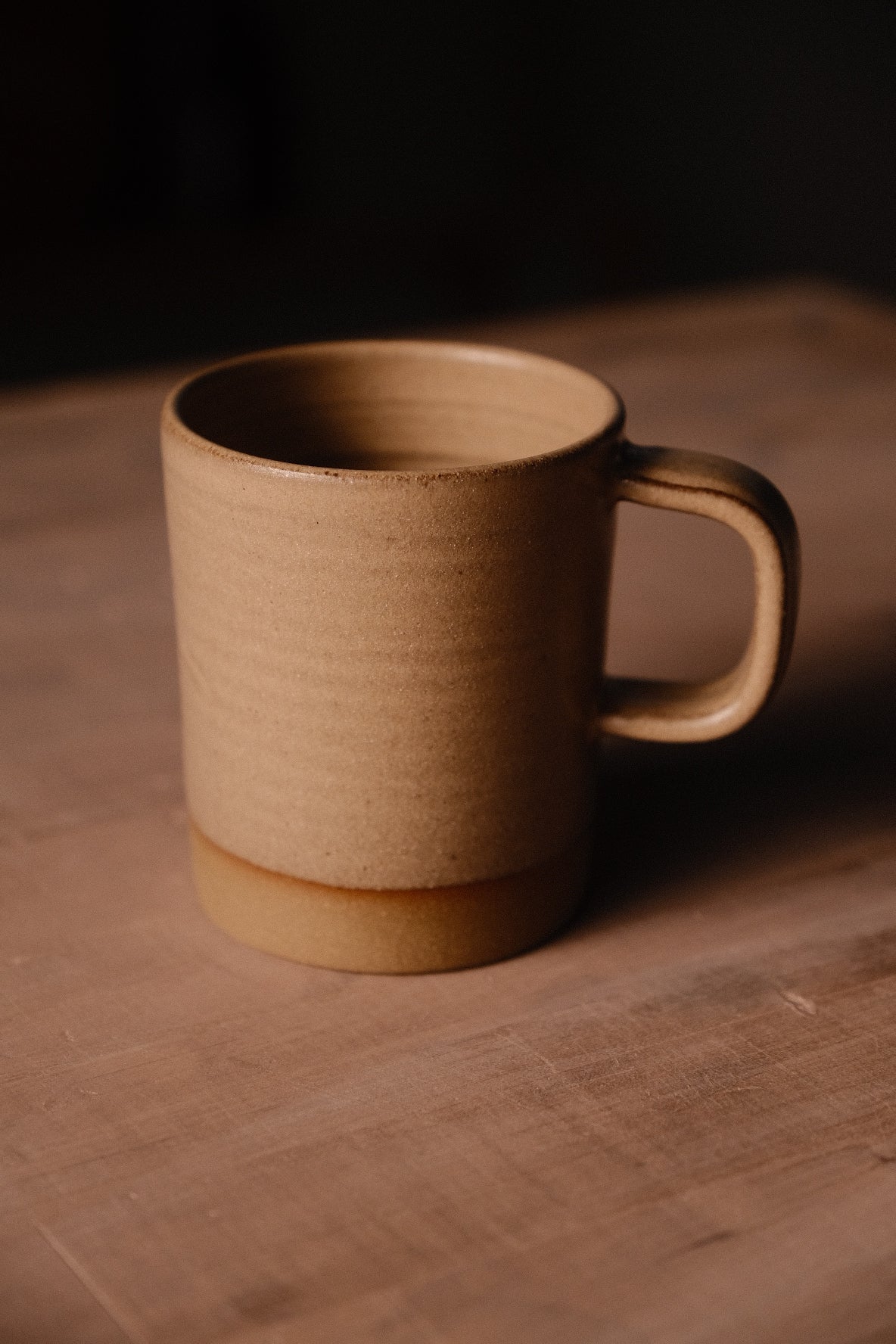 Tan Settle Ceramics Wander Cappuccino Mug on a brown tabletop, viewed at an angle to show its cylindrical ceramic body, rounded rectangular handle, fine speckles and contrasting ochre band around the base.