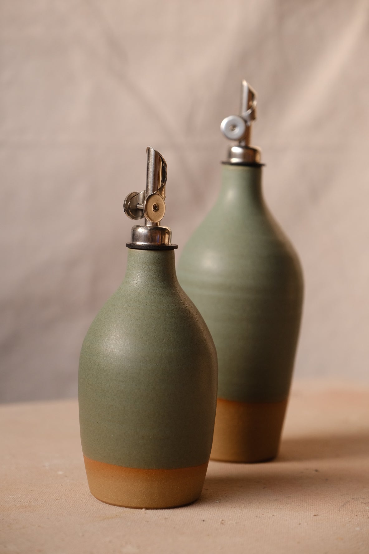 Muted olive-green Settle Ceramics Wander Oil Bottle in front of a taller sage-green bottle against beige fabric, showing fine speckles, horizontal surface rings, metal pouring spouts and ochre bases.