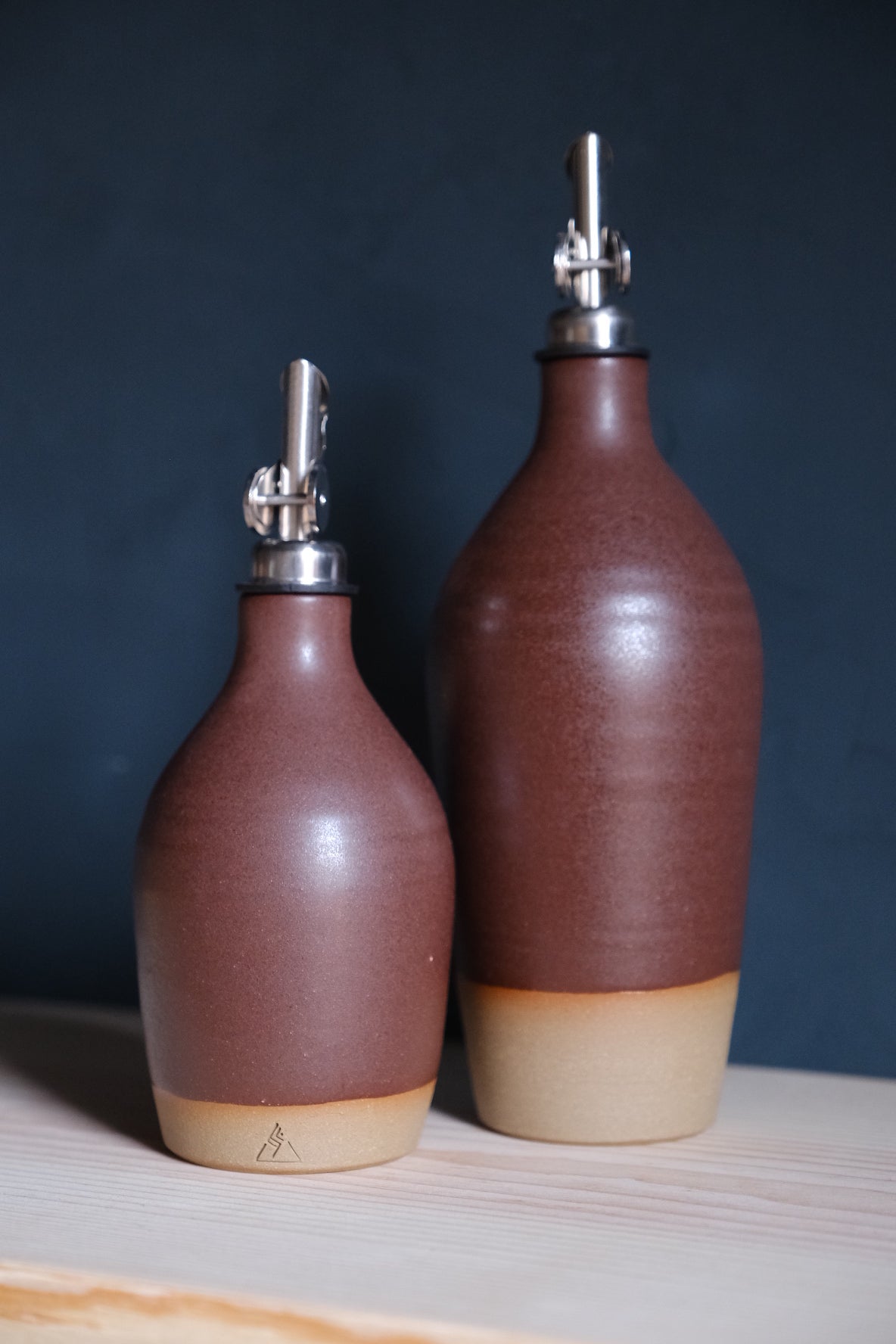 Reddish-brown Settle Ceramics Wander Oil Bottle beside a taller matching bottle against a dark blue backdrop, showing glossy ceramic bodies with horizontal rings, metal pouring spouts and sandy-beige bases.