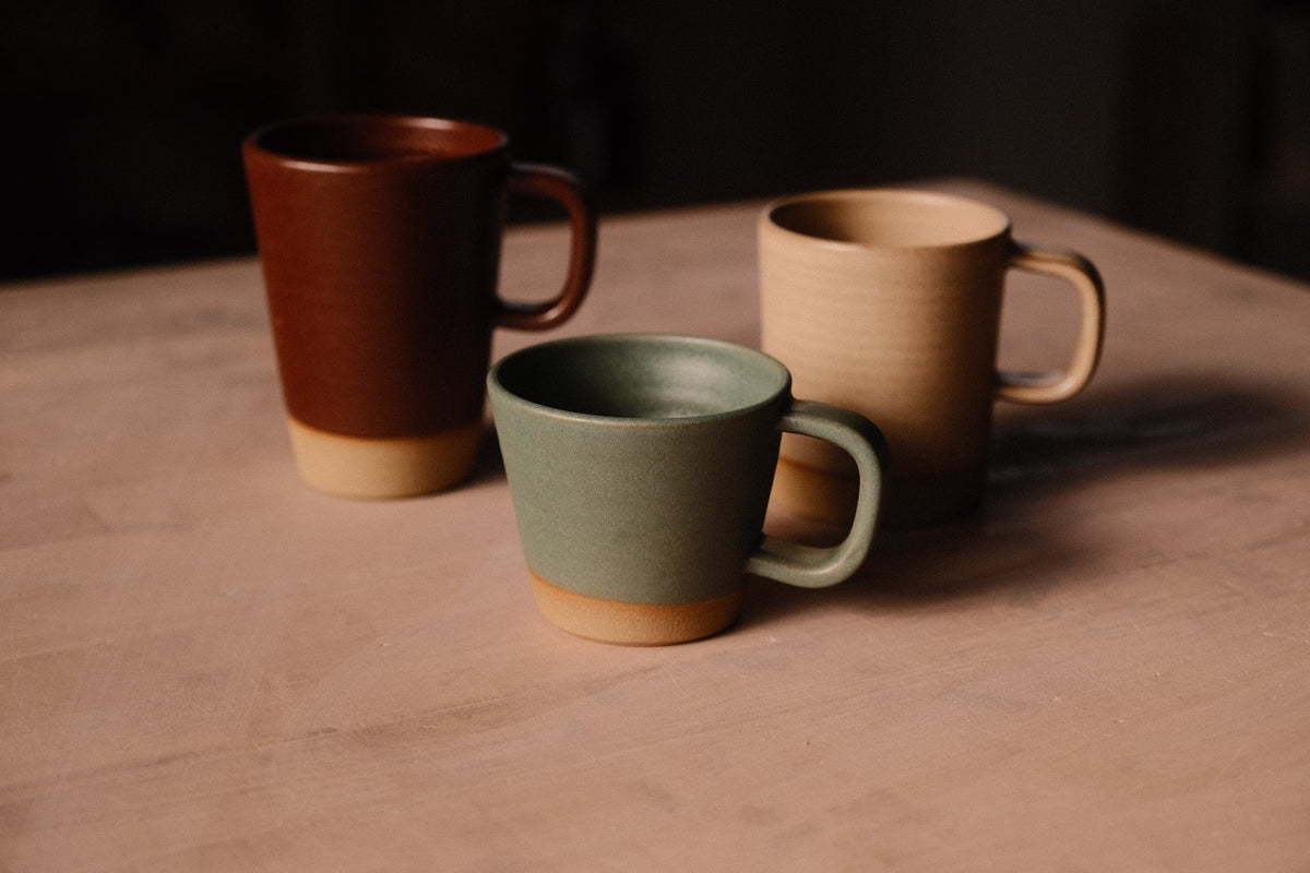 Tan Settle Ceramics Wander Cappuccino Mug beside a taller reddish-brown mug and a shorter green mug on a brown tabletop, showing their different proportions, curved handles and contrasting glaze colours.