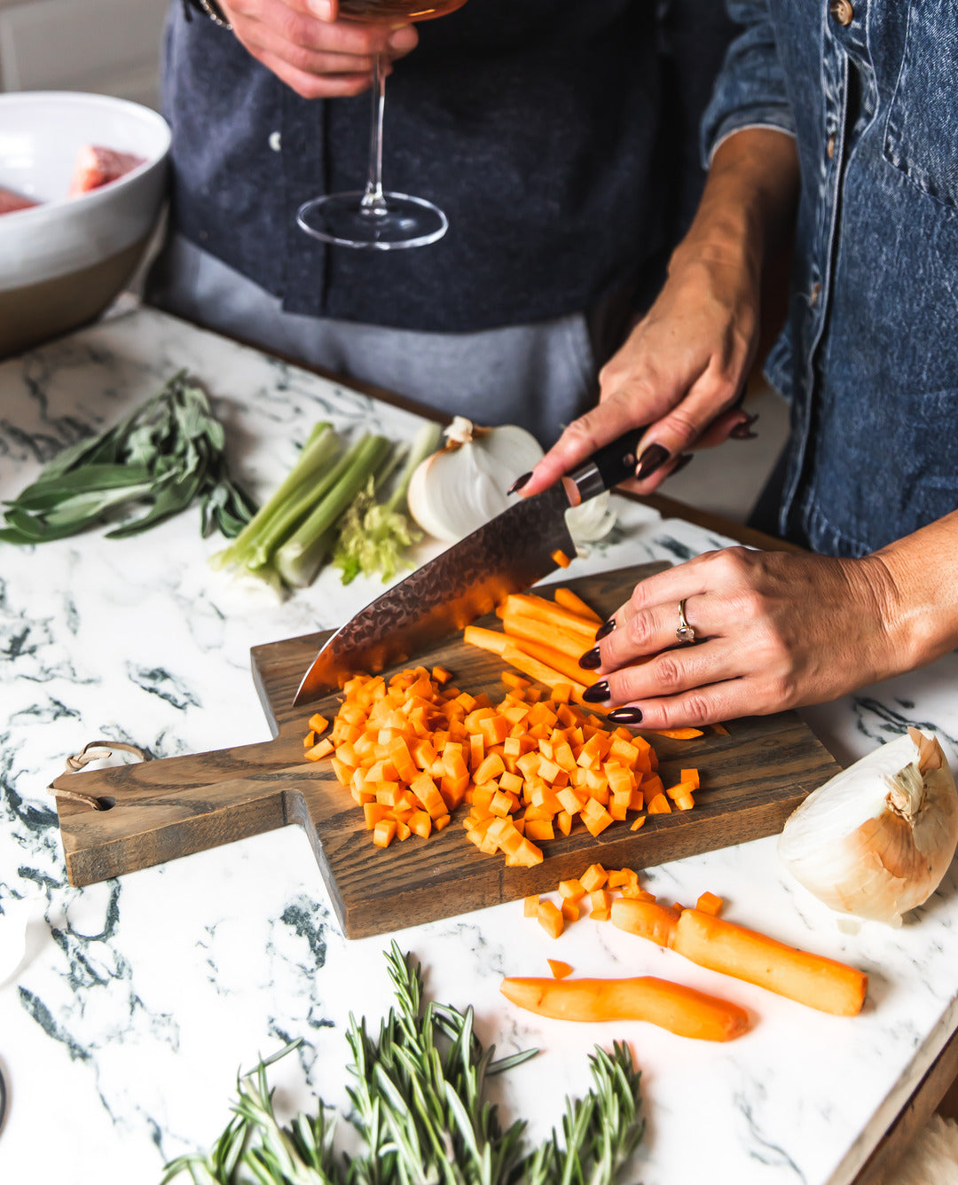 Hands chopping orange vegetables on a dark wood Pantry Cutting Board at a marble table scattered with herbs, green onions, and bread.