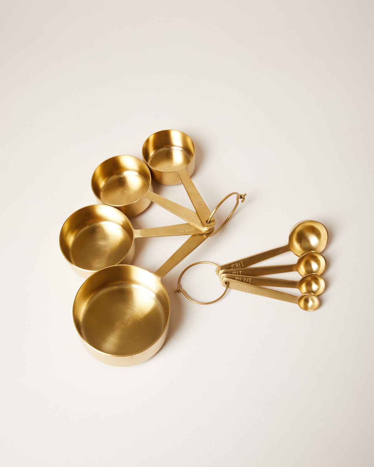 Coordinating brushed brass measuring cup and measuring spoon sets displayed side by side on a pale surface.