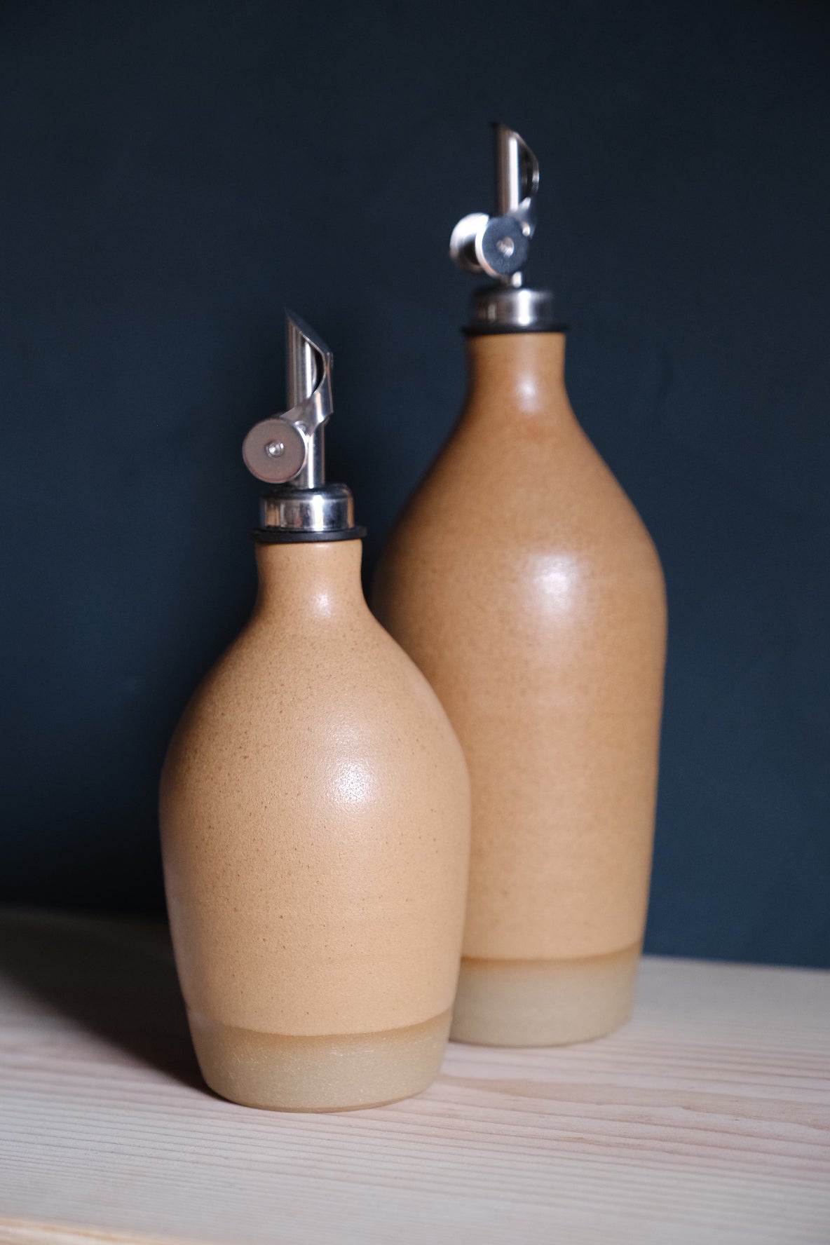 Golden-tan Settle Ceramics Wander Tall Oil Bottle beside a shorter matching bottle, showing glossy highlights, fine speckles and silver-toned pourers against a dark blue backdrop.