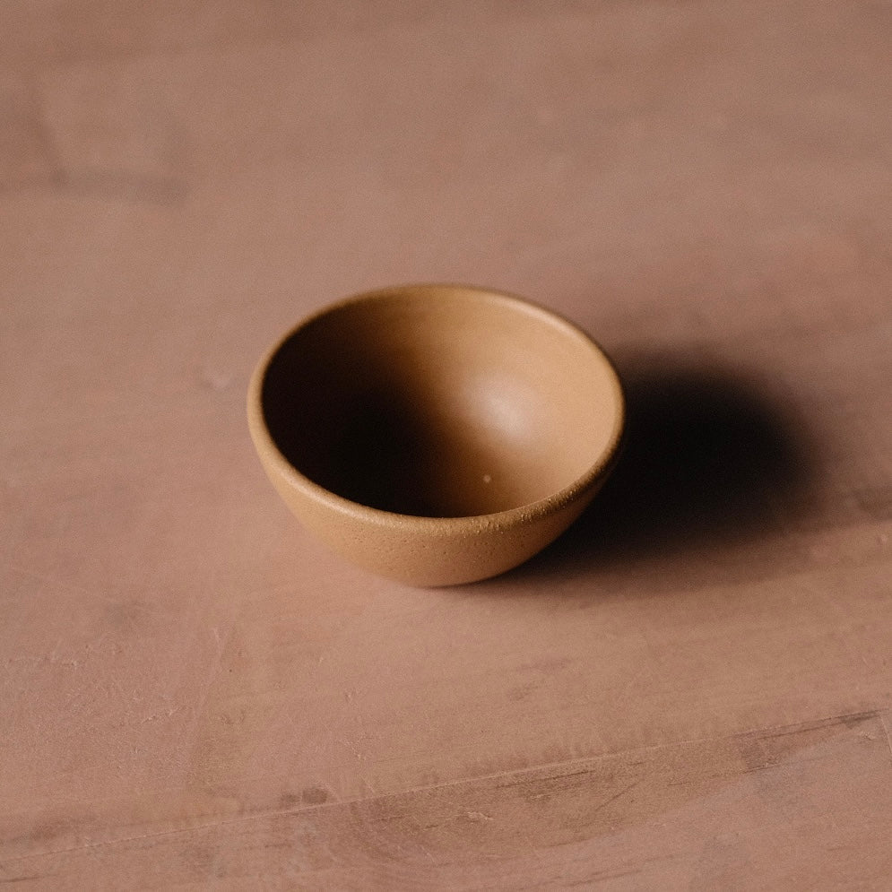 Golden-tan Settle Ceramics Wander Tiny Bowl viewed at an angle on a warm brown tabletop, showing its rounded ceramic body, gently flared rim and finely speckled glaze, with a dark shadow to the right.