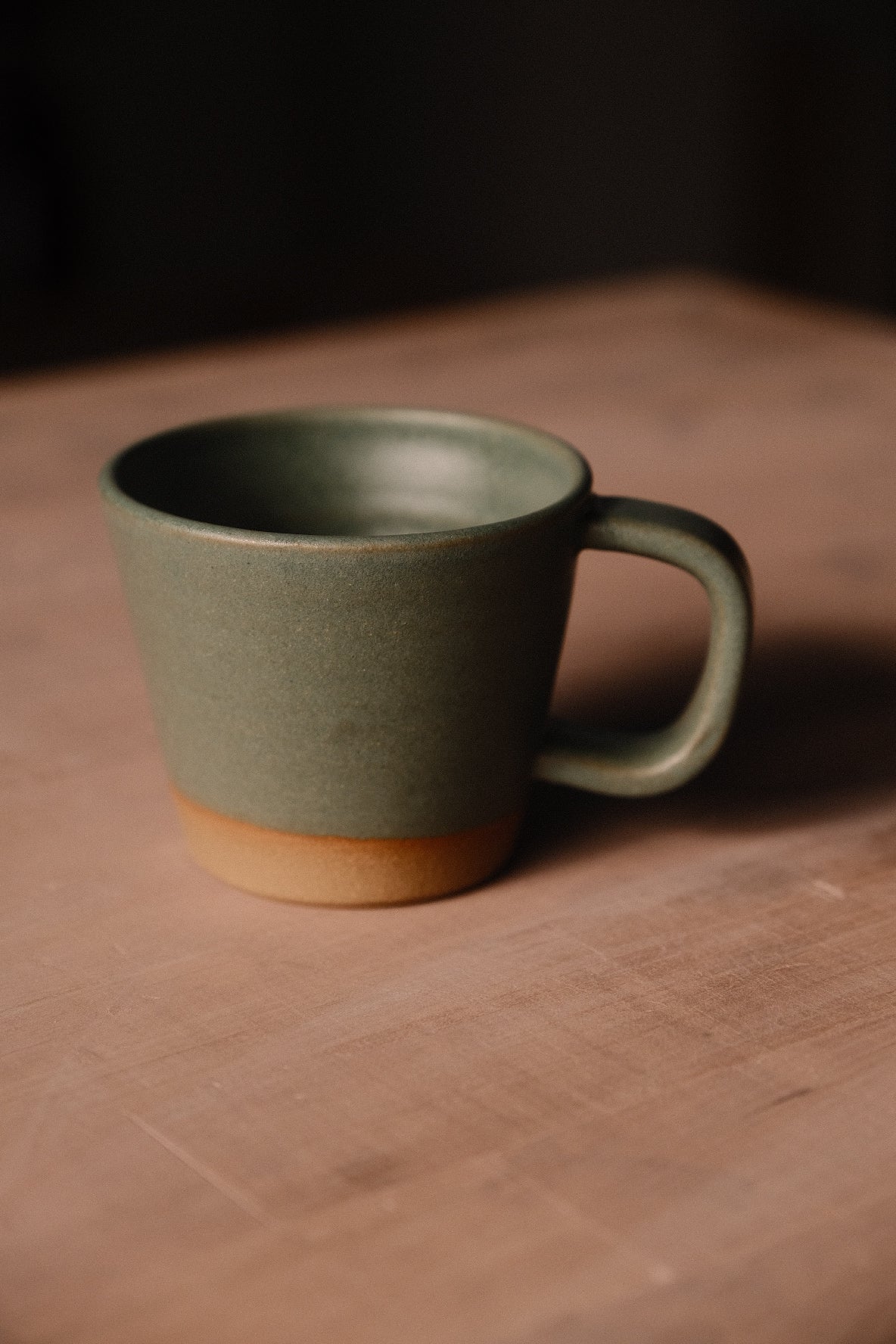 Olive-green Settle Ceramics Wander Americano Mug on a brown tabletop, showing a gently tapered ceramic body, rounded loop handle, dark glazed interior and contrasting ochre-coloured band around the base.