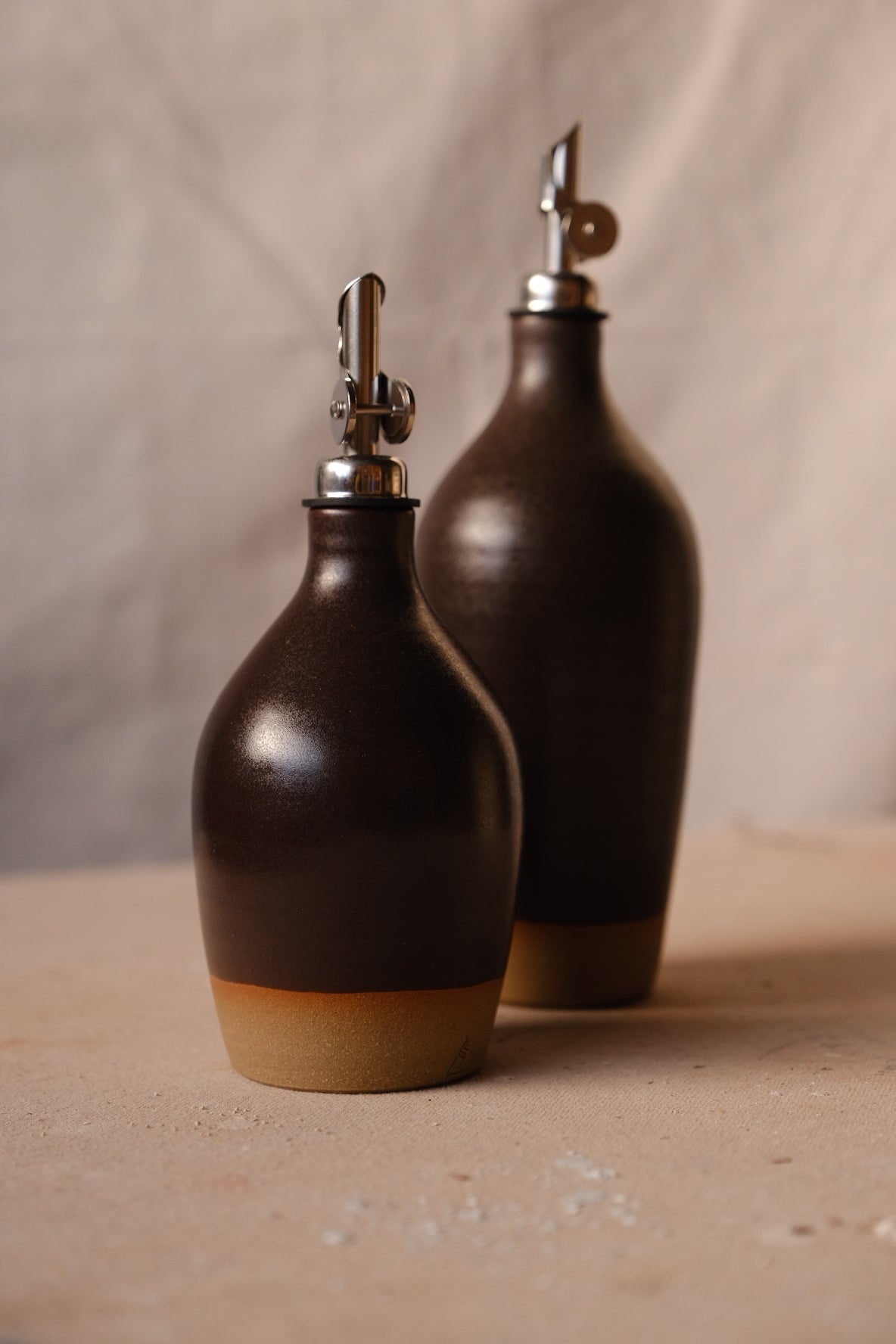 Dark chocolate-brown Settle Ceramics Wander Tall Oil Bottle behind a shorter matching bottle, with glossy rounded bodies, metal pouring spouts and contrasting ochre bases.