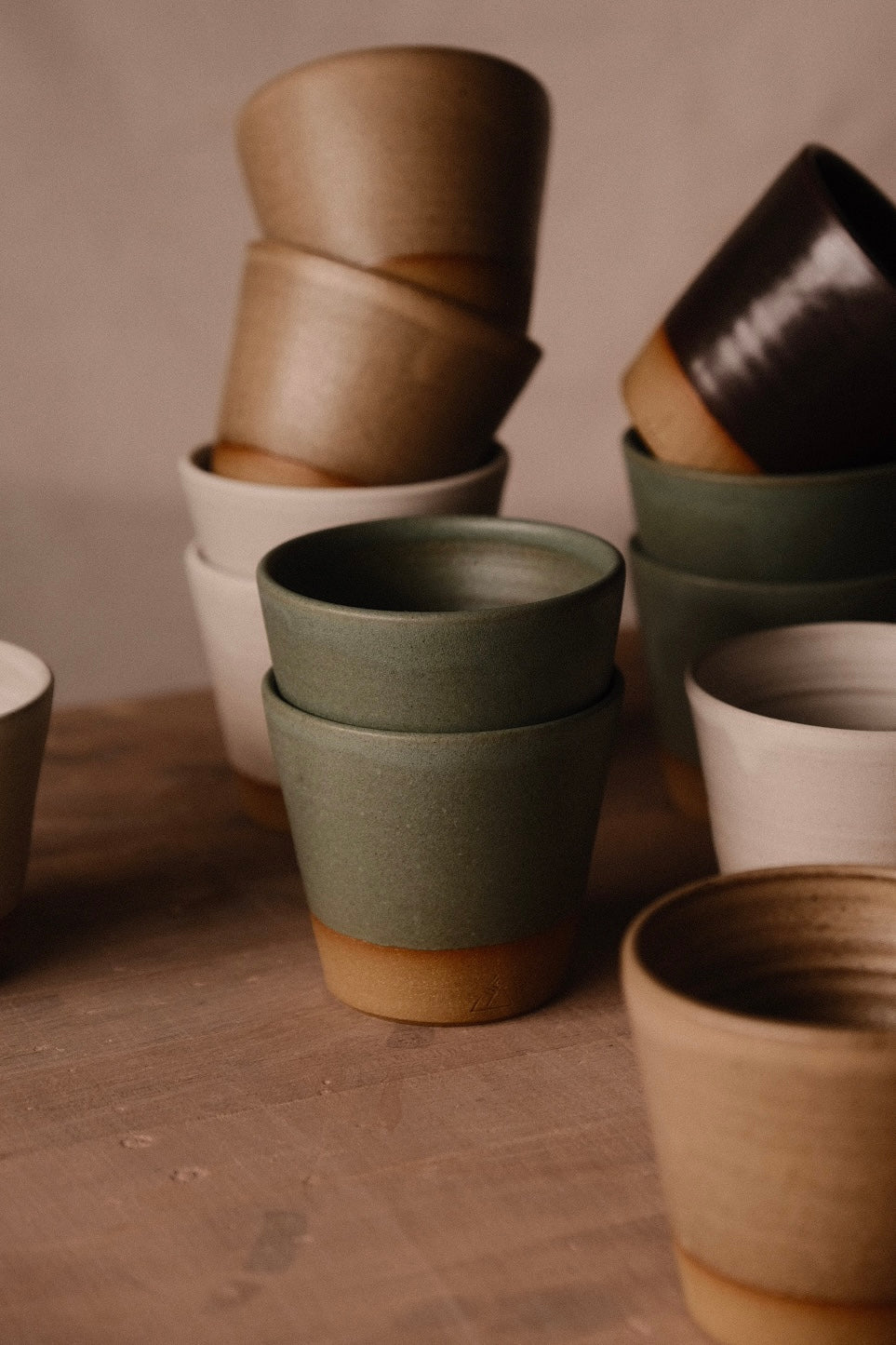 Sage-green Settle Ceramics Wander Tumblers stacked in the foreground, surrounded by cream, tan and dark brown cups on a wooden tabletop, showing handleless ceramic bodies that taper toward contrasting ochre bases.