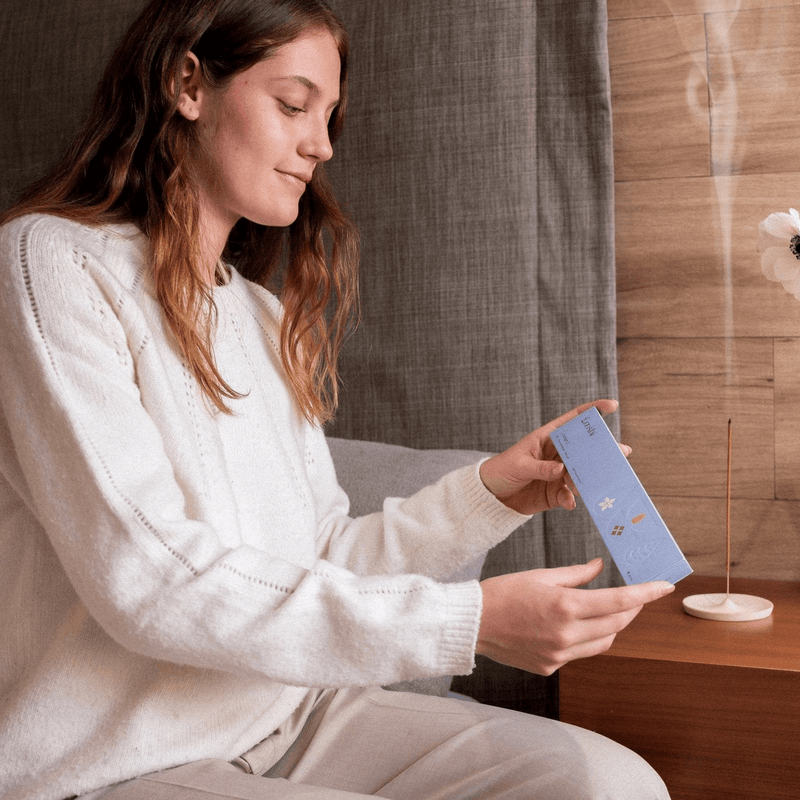 Woman seated indoors opening a light blue box of Looshi Change incense sticks beside a wooden table