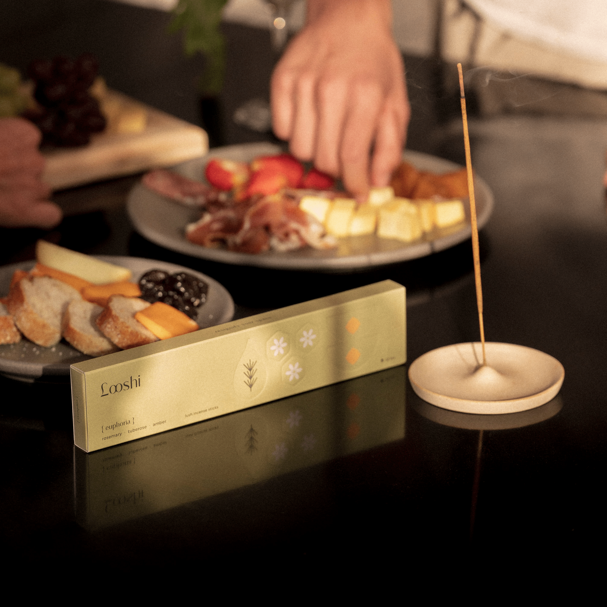 Looshi Euphoria incense box and a burning incense stick arranged beside plates of food as a person reaches across the table