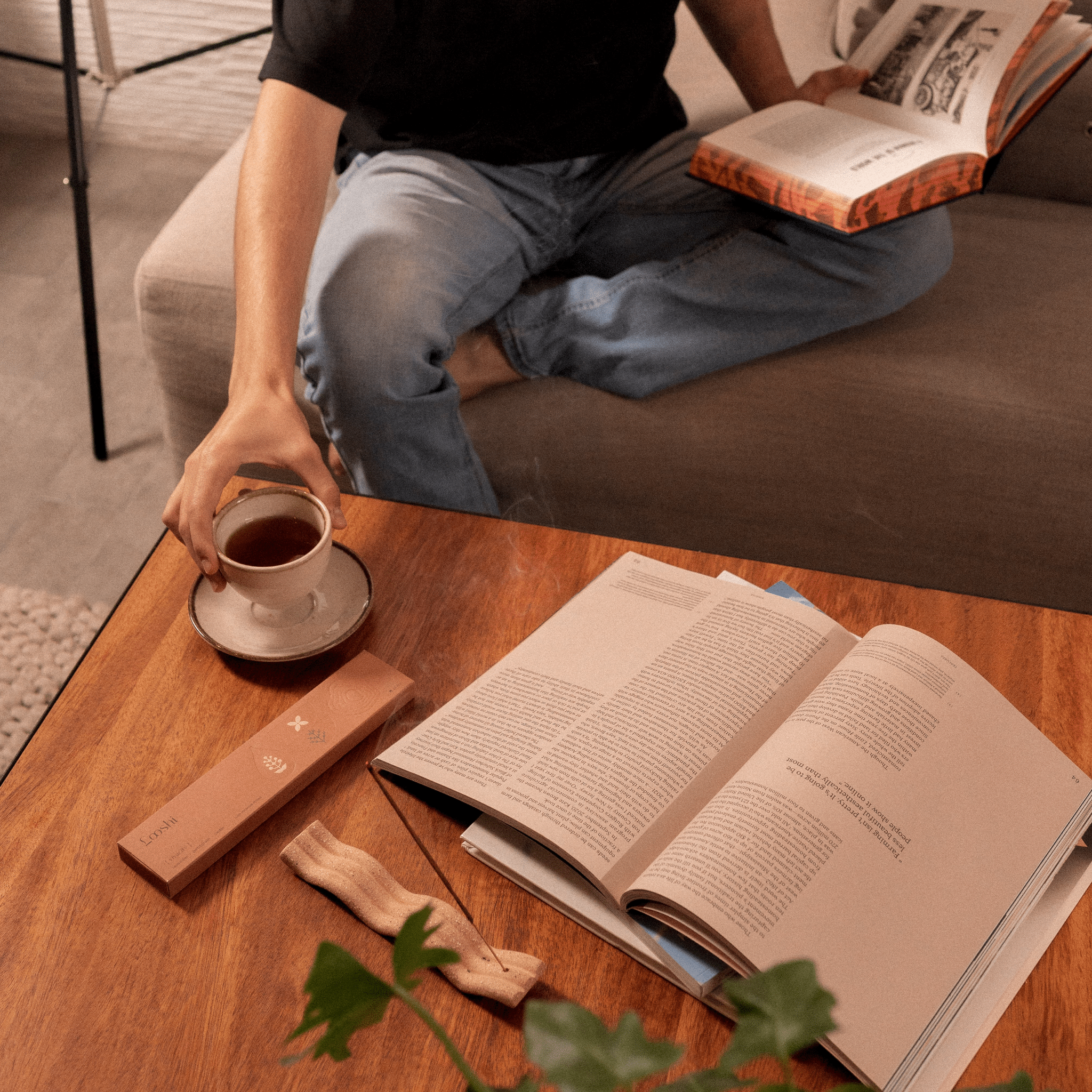 Person relaxing with a cup and an open book at a wooden table, with a box of Looshi Vivacity incense sticks and leafy sprigs nearby