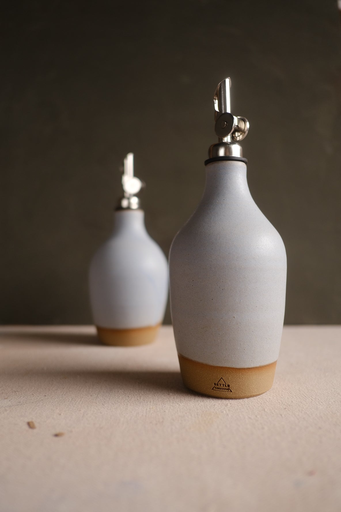 Pale grey Settle Ceramics Wander Oil Bottle in front of a smaller matching bottle on a light tabletop against a dark background, showing softly rounded shoulders, metal pouring spouts and ochre base bands.