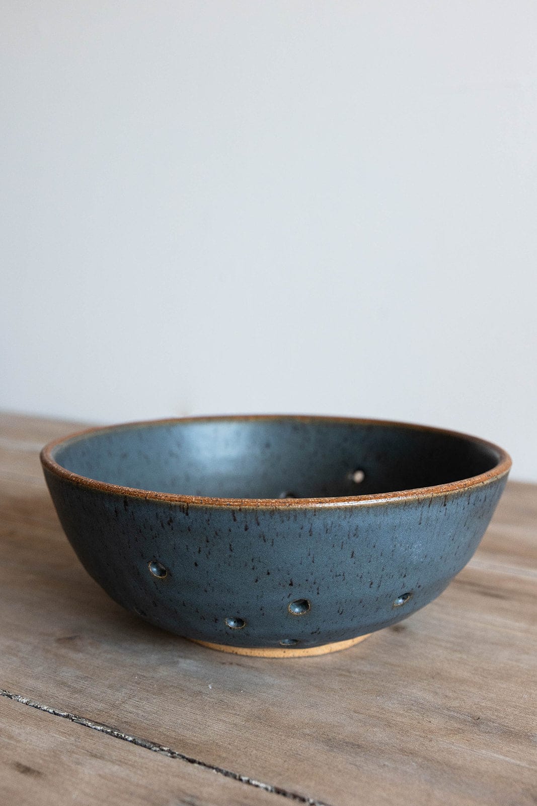 Dark blue ceramic colander bowl on a wooden surface with a light gray background
