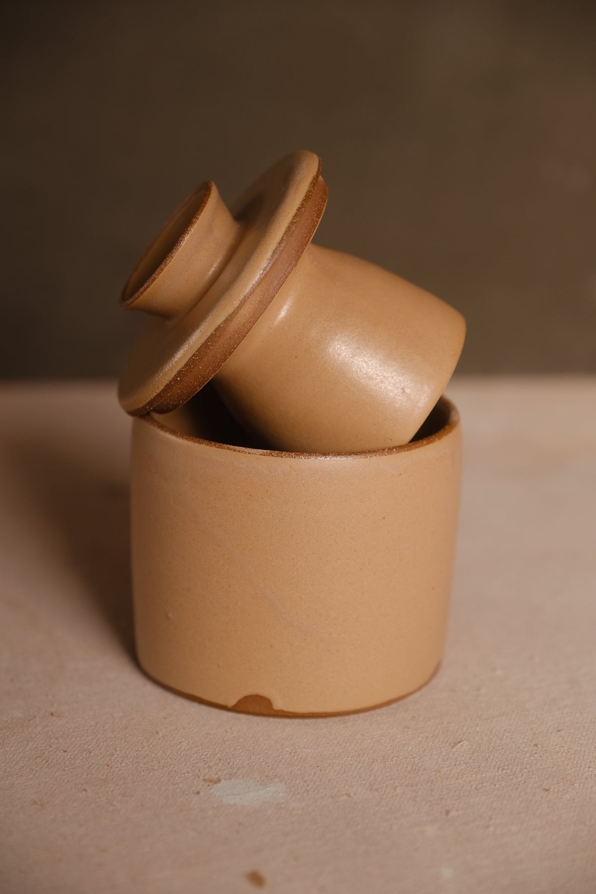 Golden-tan Settle Ceramics Butterbell with a smooth cylindrical base and matching lid tilted upward, revealing the deep ceramic cup beneath its broad rim.