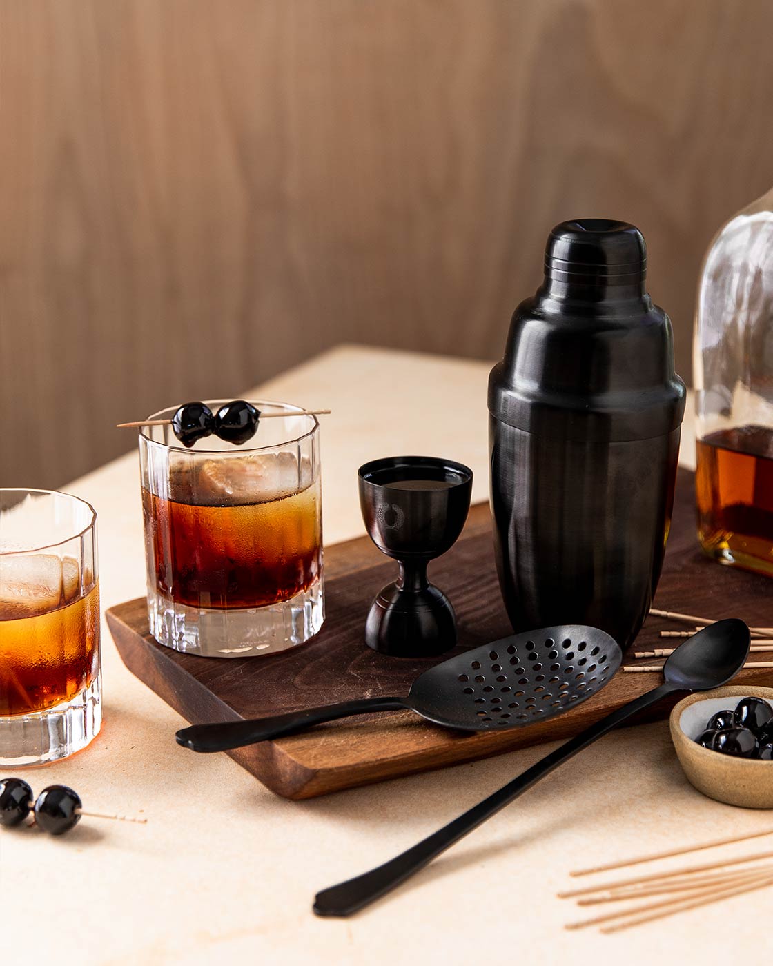 Complete brushed-onyx barware set arranged on a dark wooden board with amber cocktails, liquor bottles, blackberries, and cocktail picks.