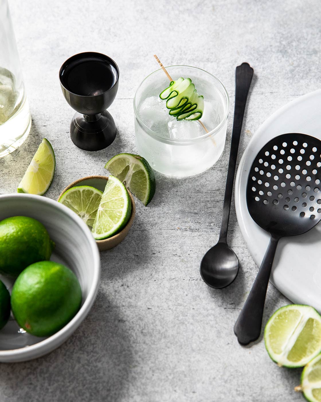 Overhead cocktail-preparation scene with the onyx jigger, strainer, and bar spoon surrounded by limes and clear drinks garnished with cucumber ribbons.