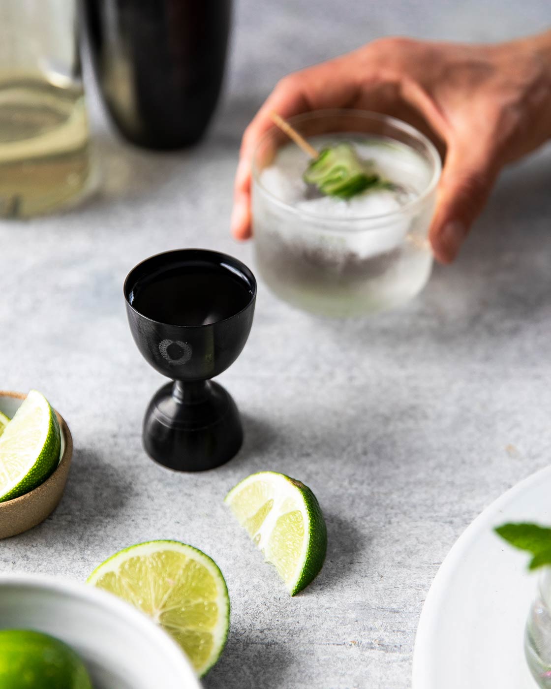 Hand placing a cucumber-garnished cocktail beside the dark onyx jigger, sliced limes, and a bowl of whole limes.