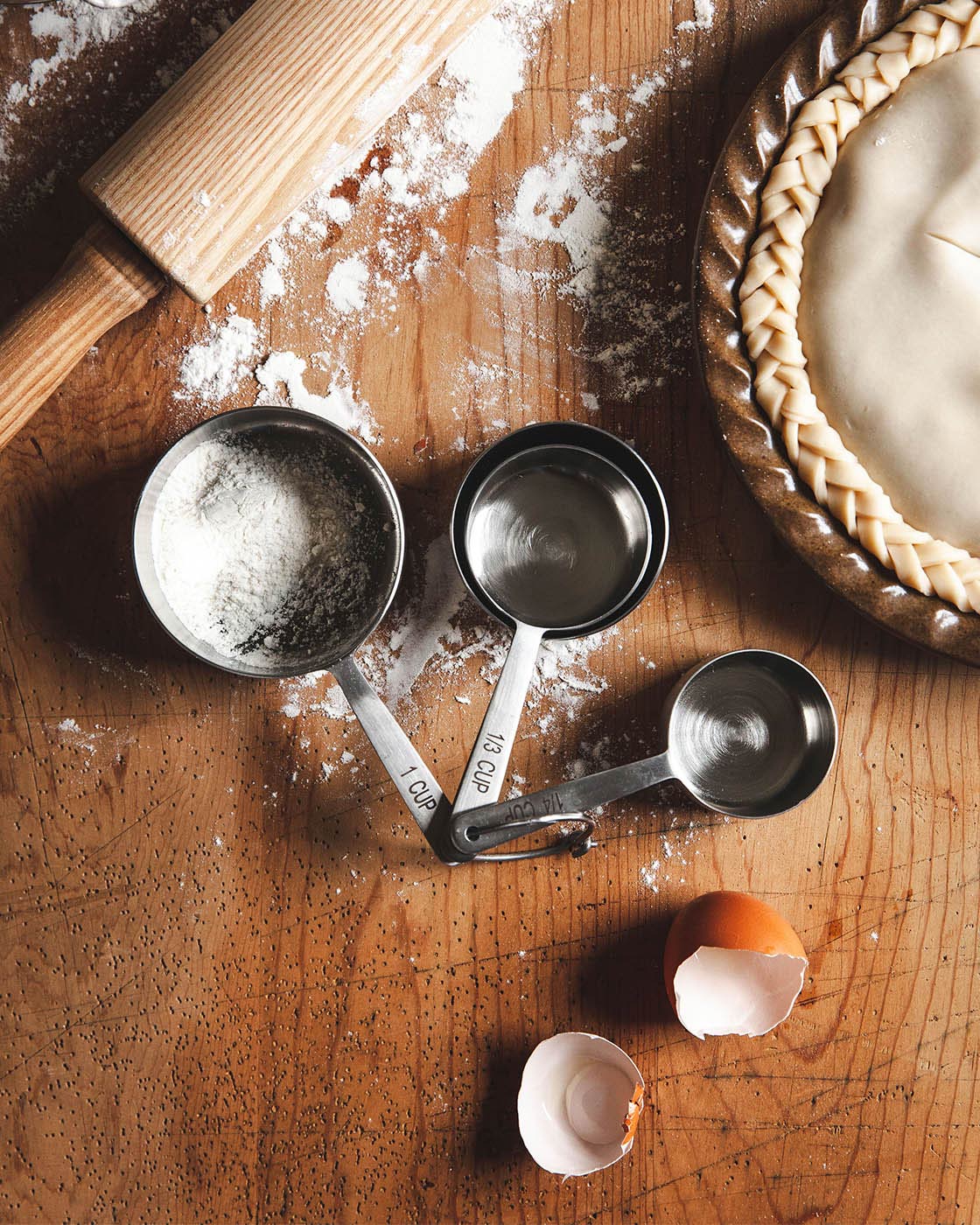 Overhead baking scene with black metal measuring cups holding flour beside a rolling pin, prepared pie crust, spilled flour, and cracked eggshells.