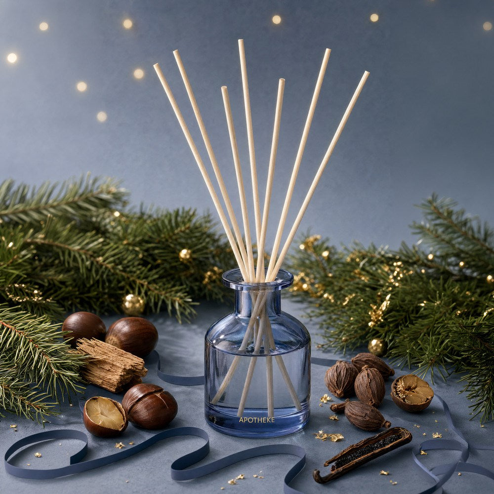 Blue-tinted Cardamom Chestnut diffuser surrounded by evergreen branches, chestnuts, wood pieces, and spices on a blue surface, with curled ribbon, scattered gold accents, and warm lights behind.