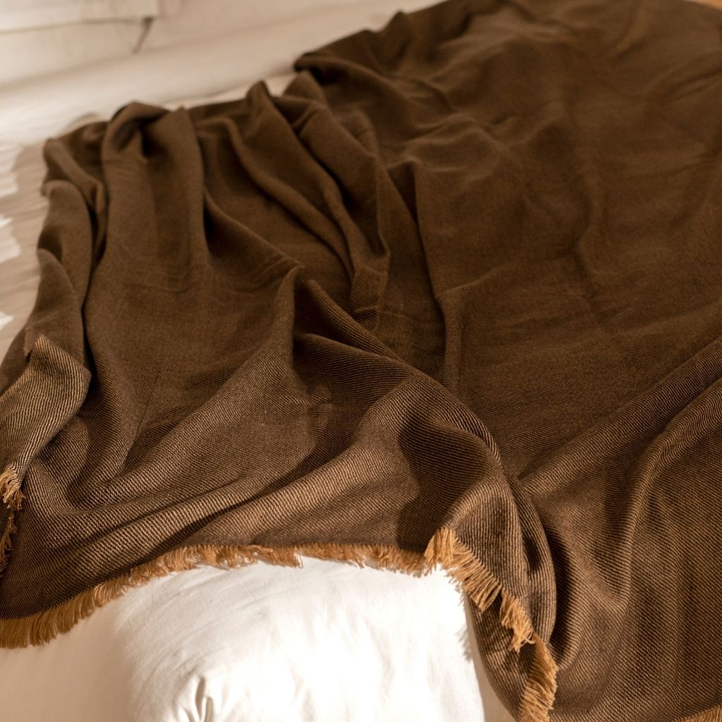 Hay Brown merino throw loosely gathered across white bedding, showing its soft fluid drape and fringed border