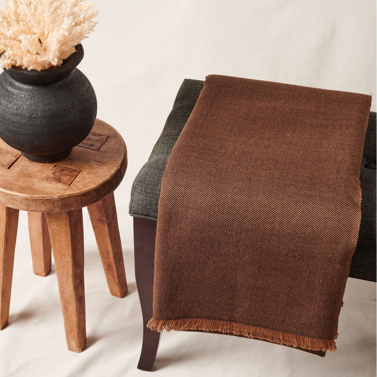 Variously Hay Brown merino handwoven throw draped over a charcoal chair beside a rustic wood stool with a black vase and dried grasses