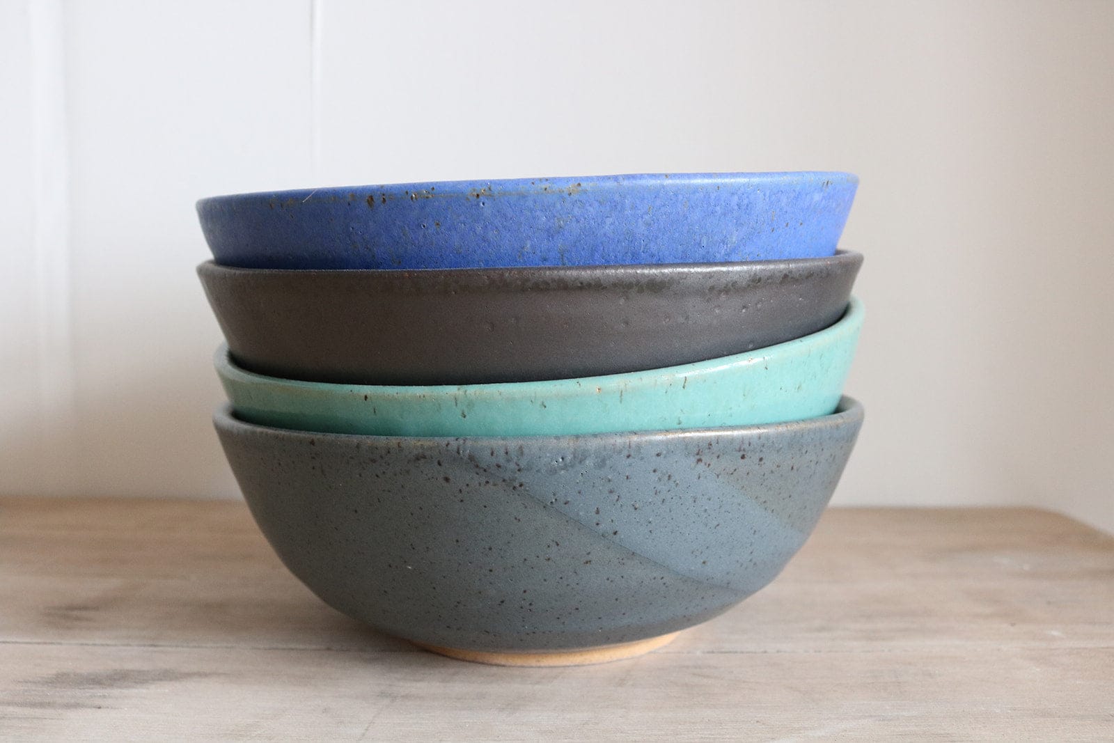 Stack of four ceramic ramen bowls in blue, gray, and green on a wooden surface.
