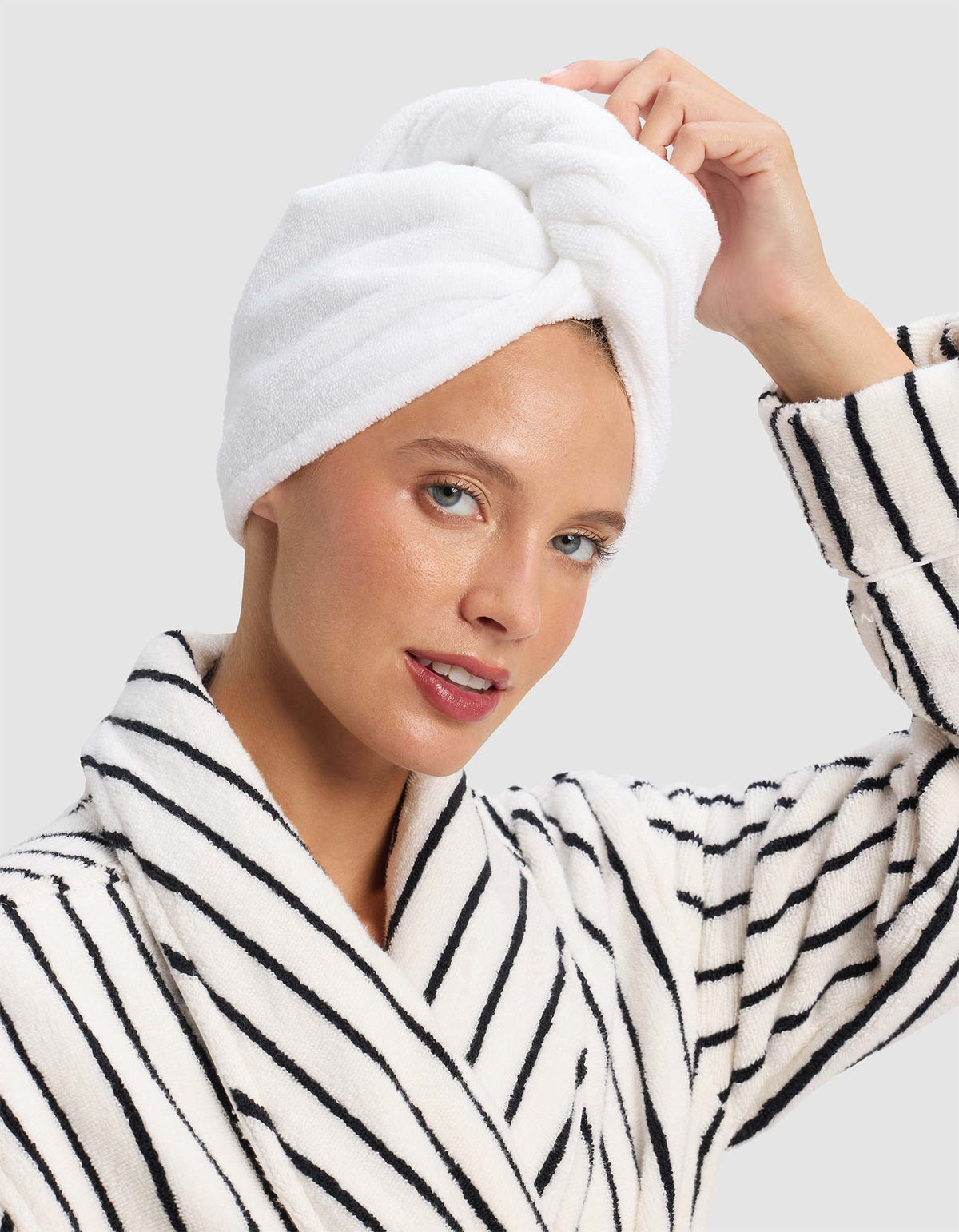 Model adjusting a white Luxe Hair Towel twisted into a wrap on top of her head, wearing a white robe with narrow black stripes.