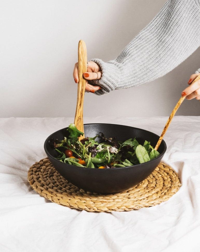 Person holding a black bowl with salad on a woven mat, The Salad Essential brings together Salad Bowl and Forks.