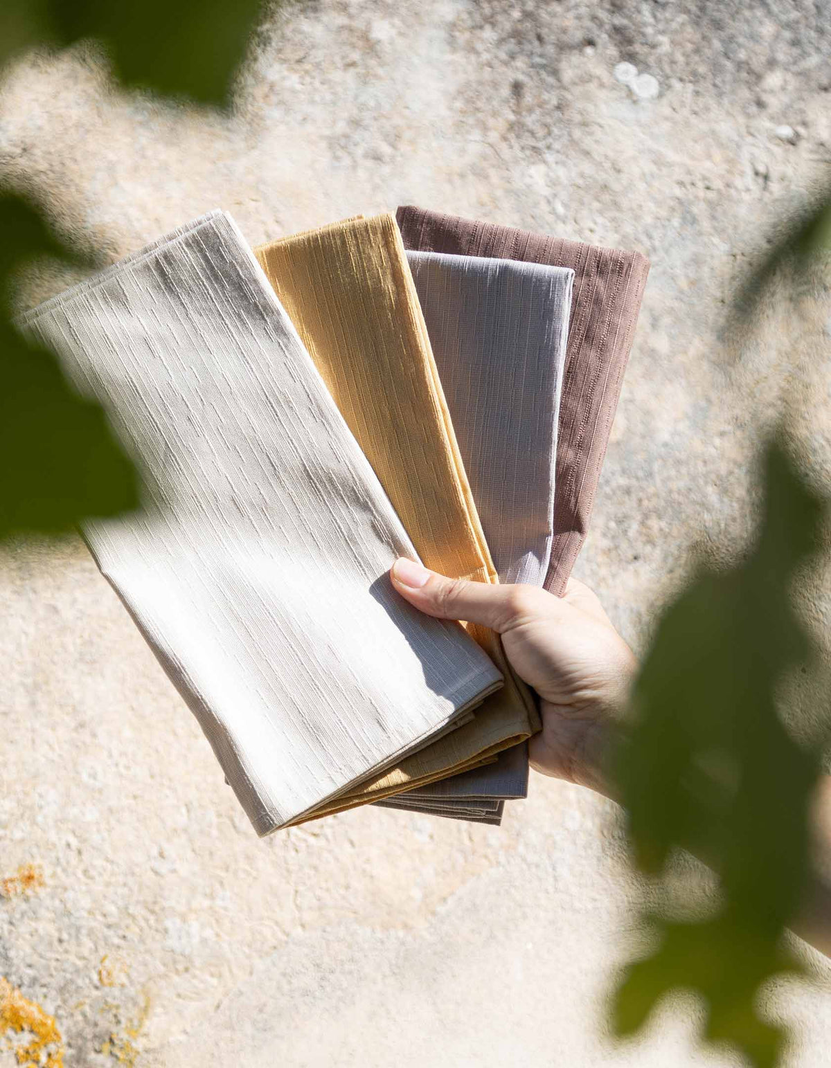 A hand holding fanned Tortuga Forma Dinner Napkins Sabi Mixed Sets in pale grey, mustard yellow, muted mauve and charcoal-grey tones outdoors, with sunlight and leafy shadows falling across a rough stone background.